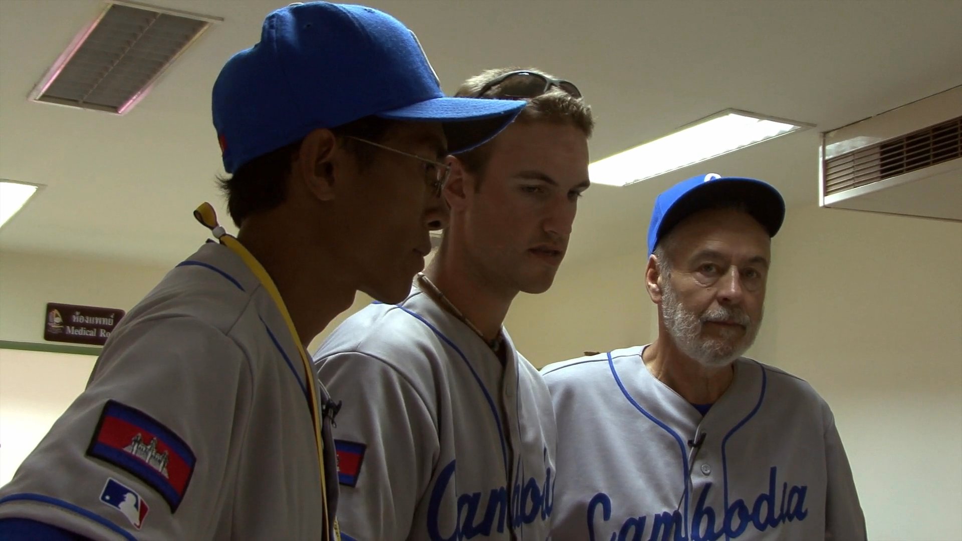 A coach provides guidance and instruction to Cambodian national baseball team players during a meeting in a locker room setting. The athletes listen attentively while seated and standing as they prepare for competition.