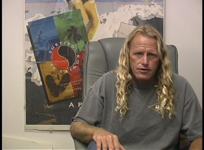 A long-haired professional surfer sits in an office setting, gesturing while delivering an interview or commentary.