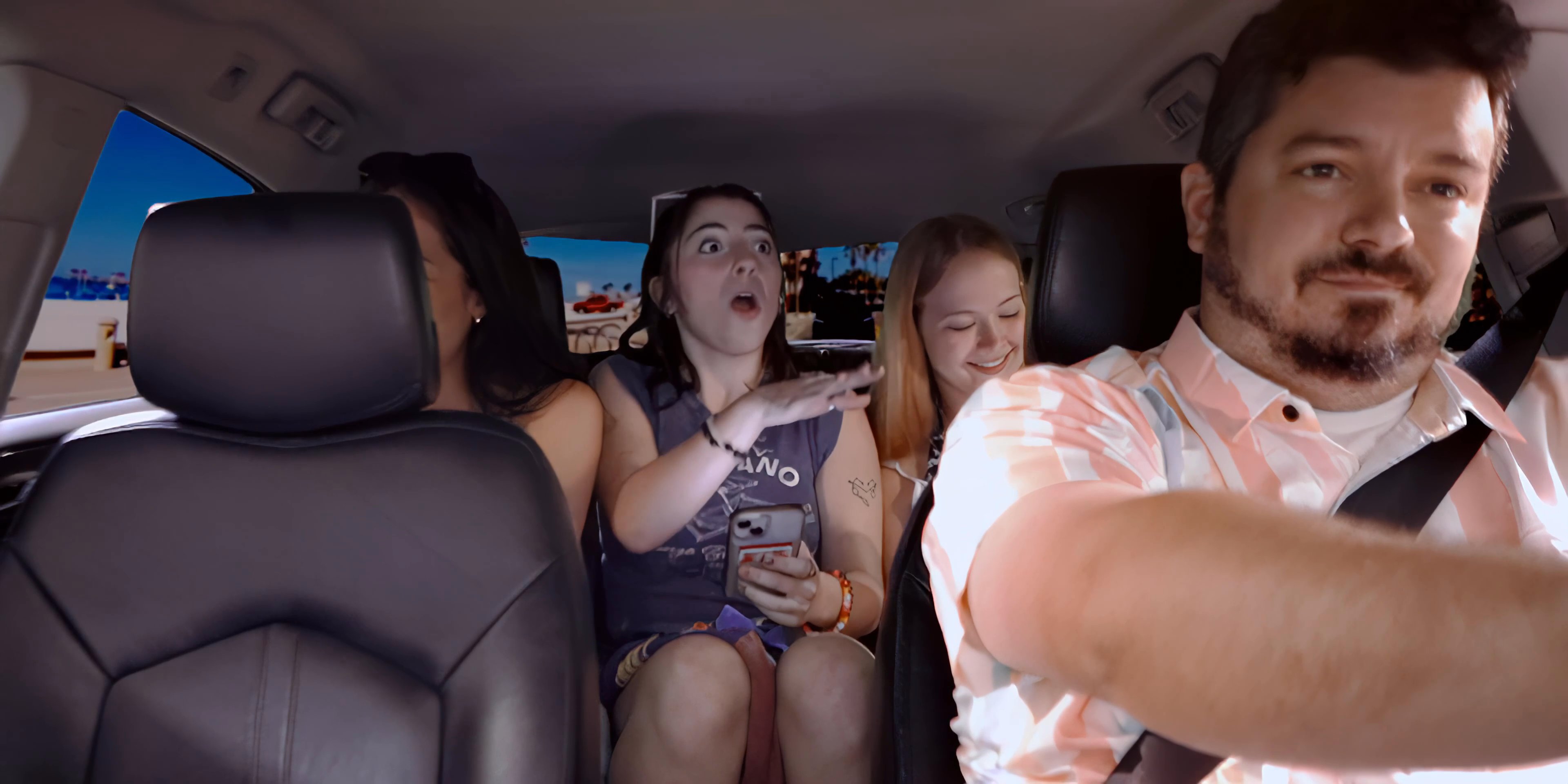 A father drives his daughters in a car while engaged in an animated group conversation. The family members express varied reactions and emotions during their commute.