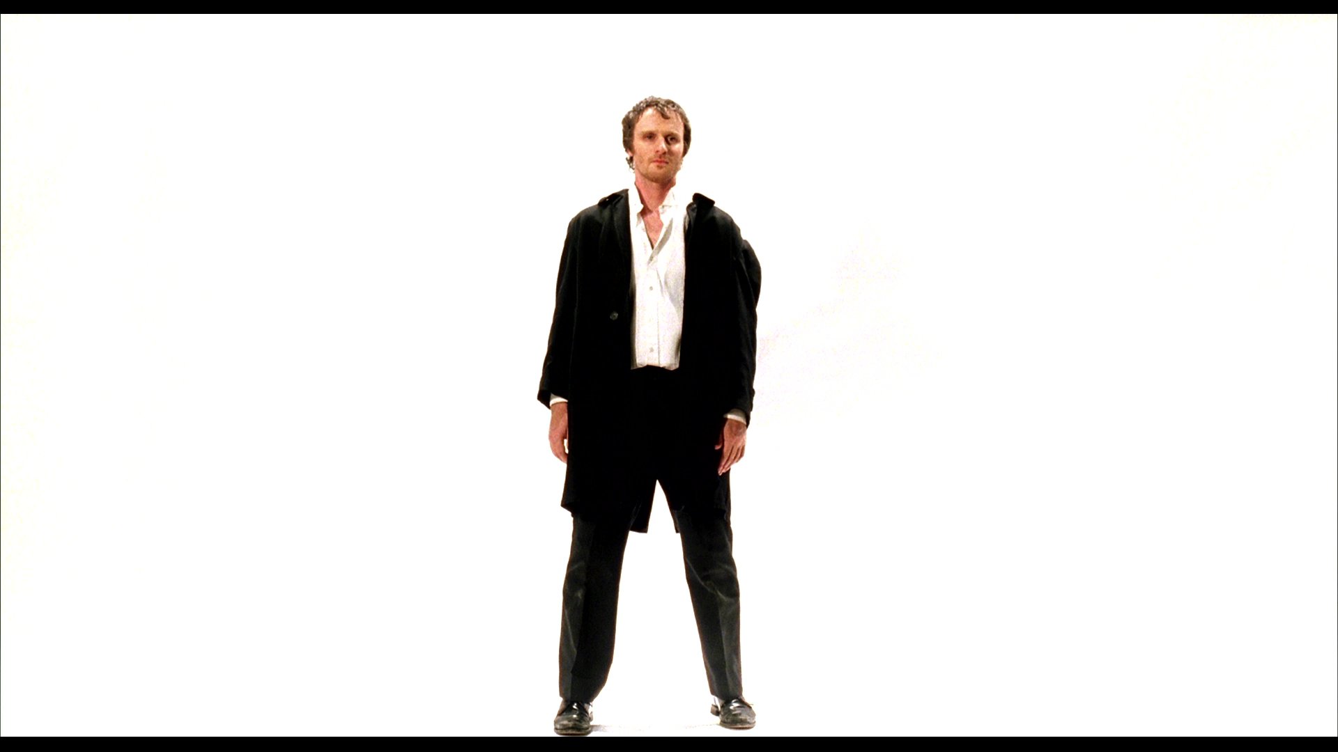 A man in a dark coat performs a series of rhythmic, stylized movements against a stark white background in a minimalist art film. The sequence follows a strict set of constraints highlighting controlled physical expression.