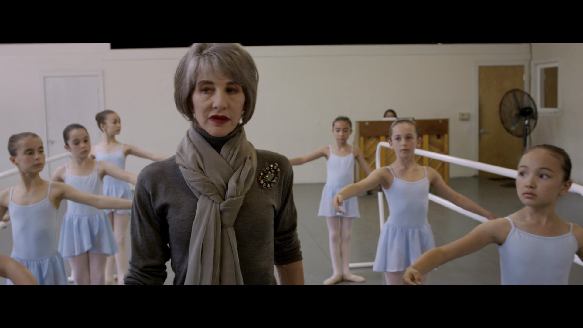A professional instructor guides a group of young ballet students through technique training in a studio setting. The session focuses on precision and proper form at the ballet barre.