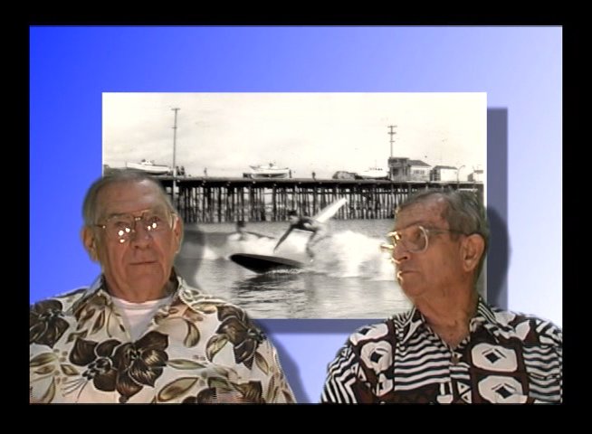Two men discuss historical memories against a backdrop of archival photographs showing coastal scenes and piers. The conversation emphasizes personal recollection and cultural history.