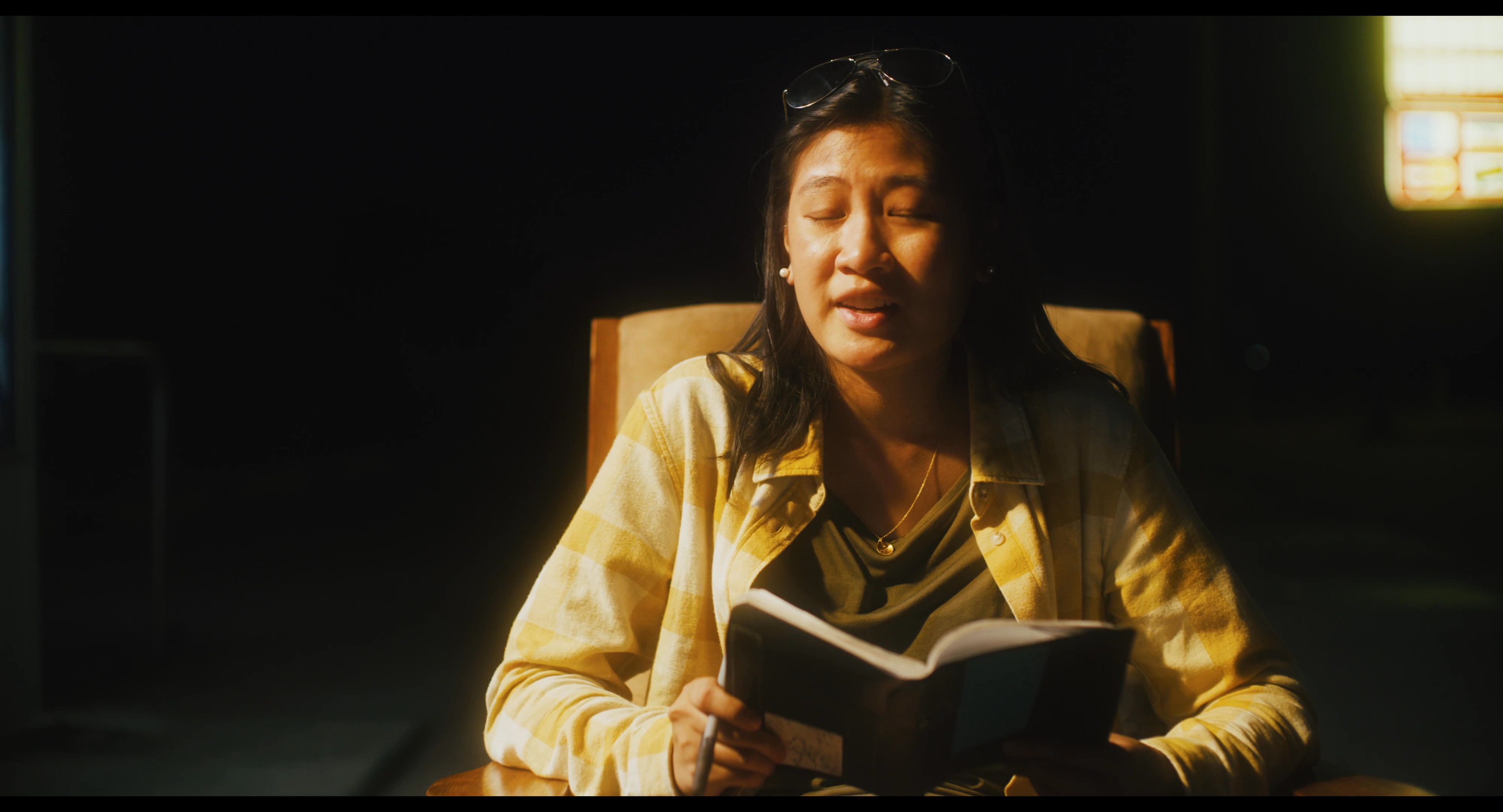 A woman reads a book while sitting in a rocking chair outside a storefront, engaging in conversation with a man seated nearby during the evening.