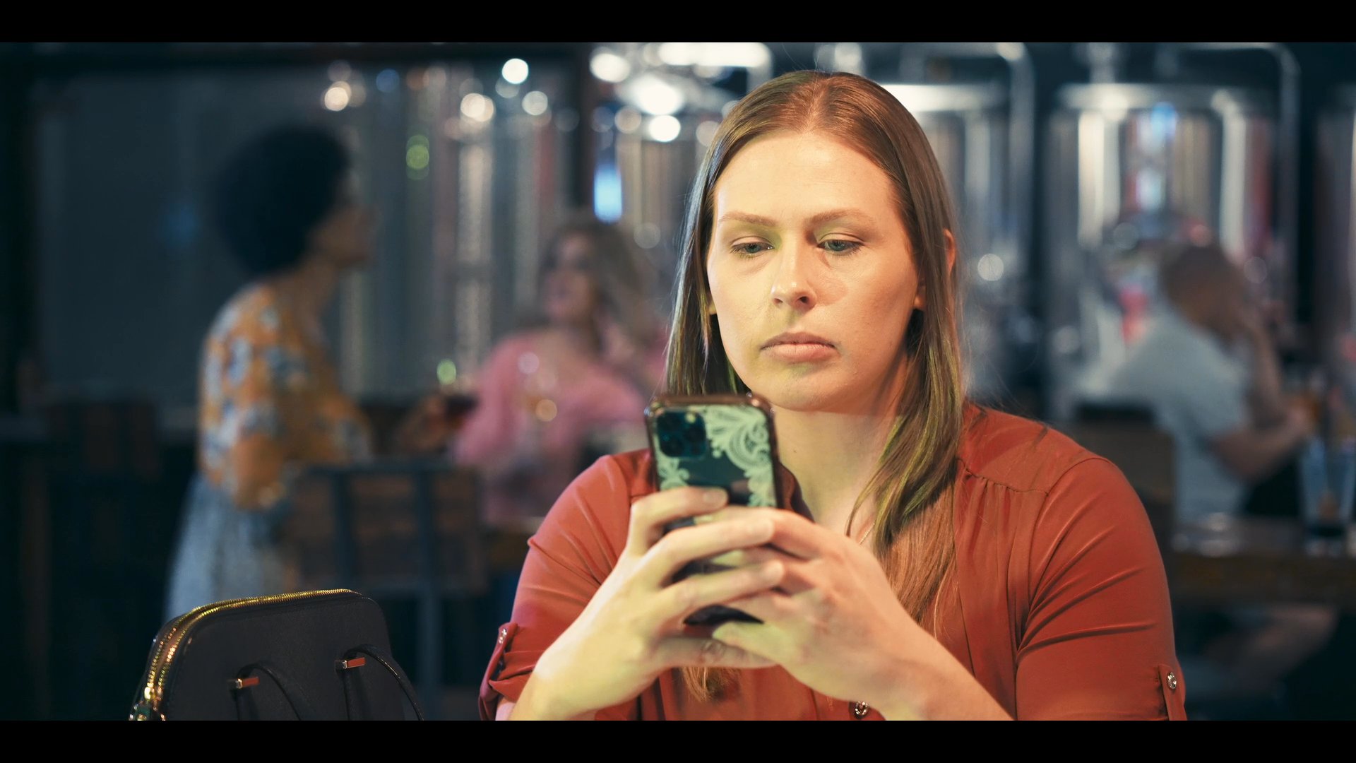 Patrons enjoy beverages and conversation in a brewery setting while a woman sits at a table focusing on her smartphone.