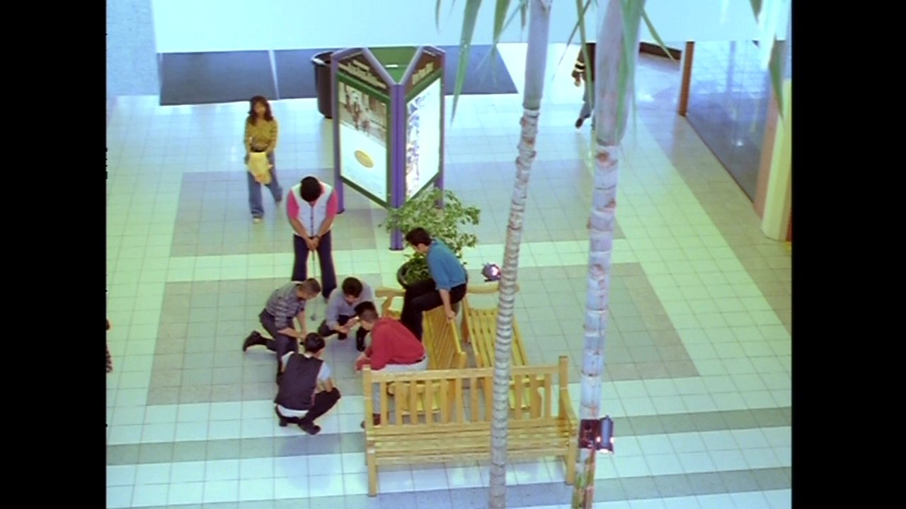 Individuals navigate and congregate within a bustling shopping mall, interacting near a music store and central seating area.