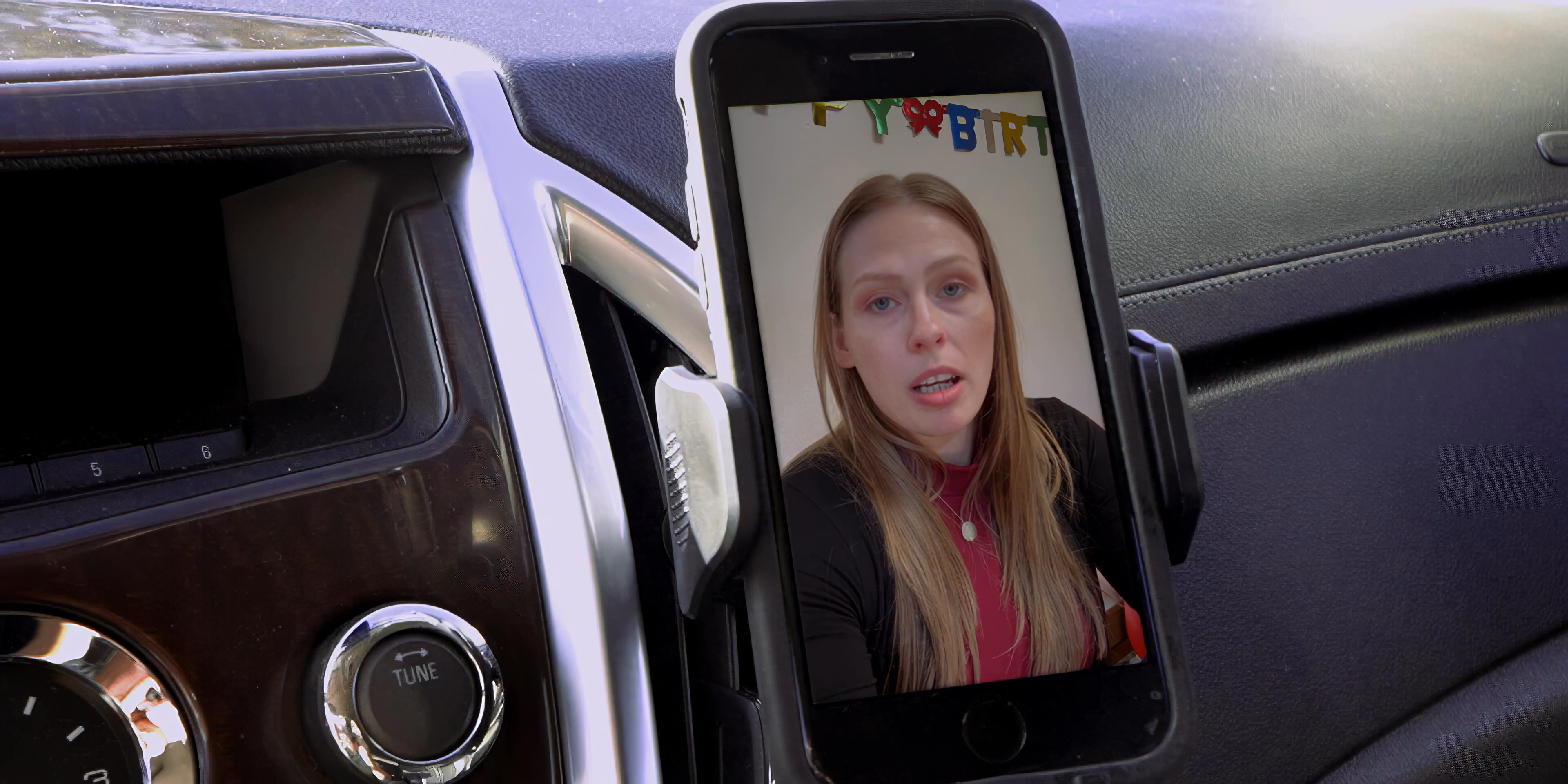 A man in a vehicle engages in a video conference on his mounted smartphone while parked near a palm-lined beach.