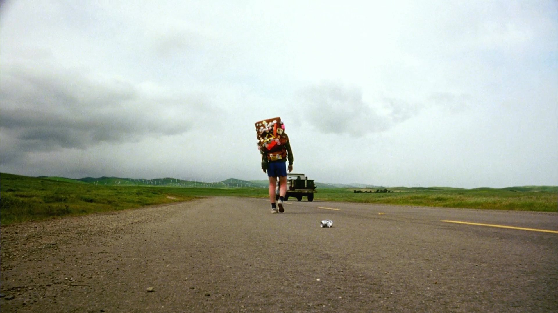 A backpacker trekking along an open rural road drops an object and stops to retrieve it while a vintage truck passes by. The traveler continues their journey across the vast, grassy landscape under an overcast sky.