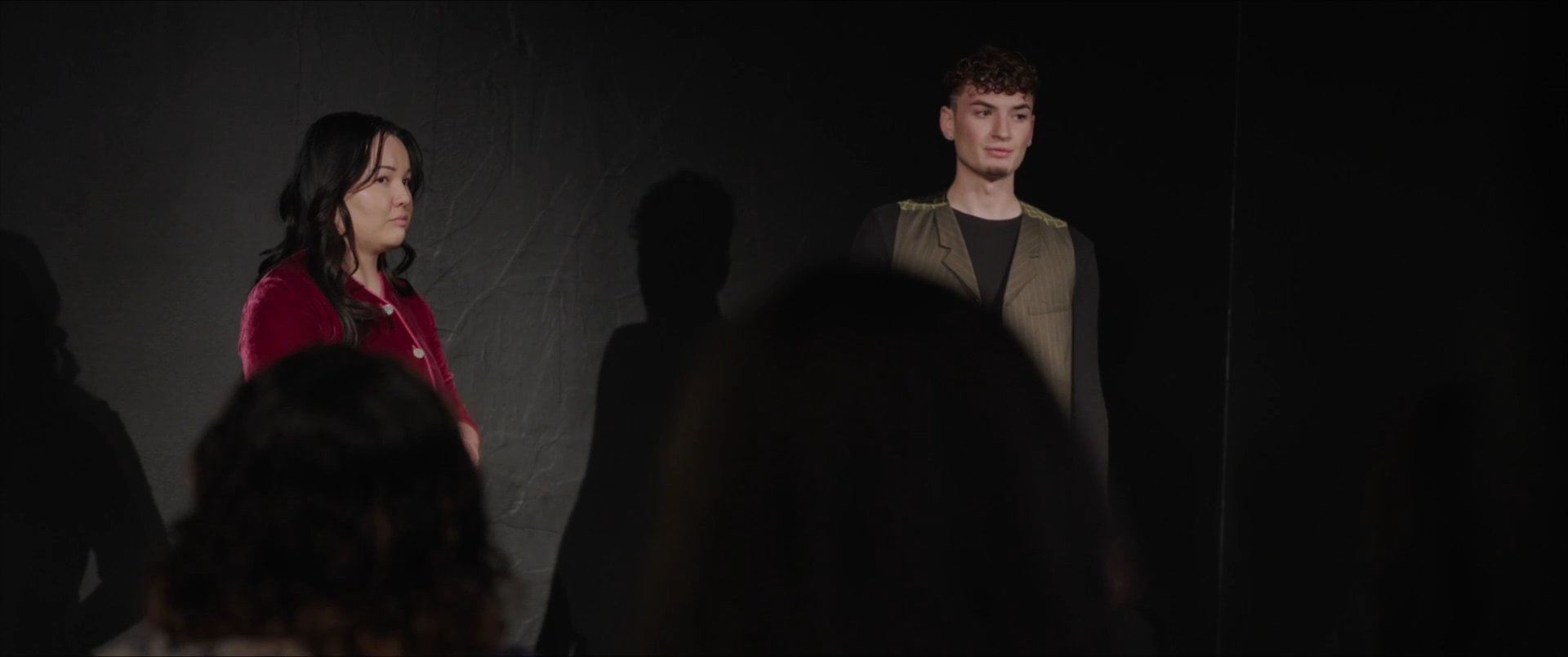 Actors in costume perform a scene on stage during a rehearsal session while others watch from the audience area. The performance shifts between dramatic dialogue and focused character interaction under theatrical lighting.