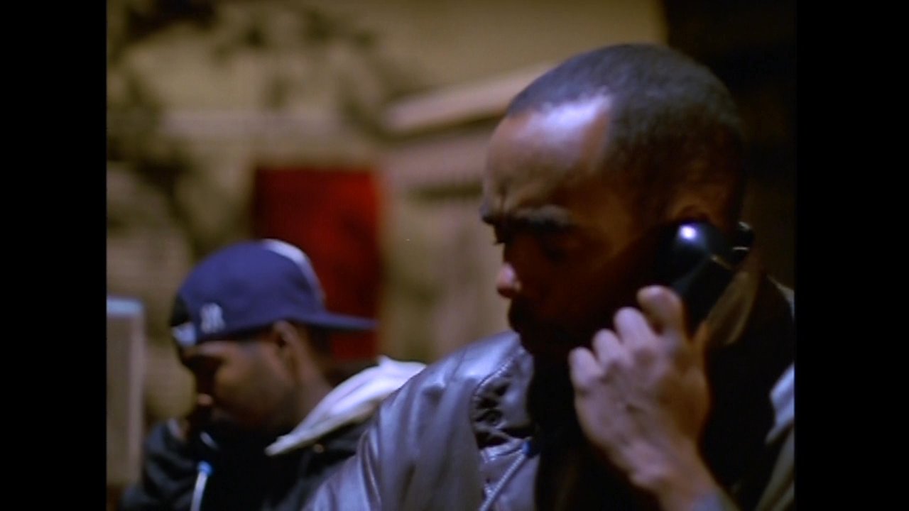 A man in a leather jacket initiates a telephone conversation at an outdoor public phone booth during the evening hours. He monitors his surroundings before focusing on the call.