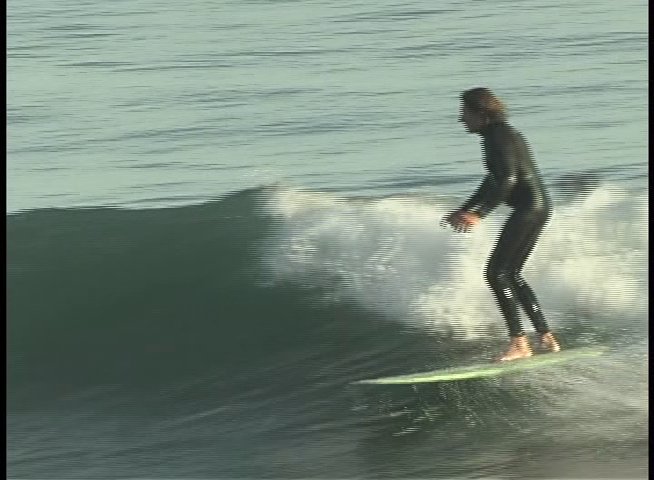 A man in a wetsuit skillfully maneuvers a surfboard across breaking ocean waves. The sequence alternates between close-up commentary and action shots of the surfing performance.