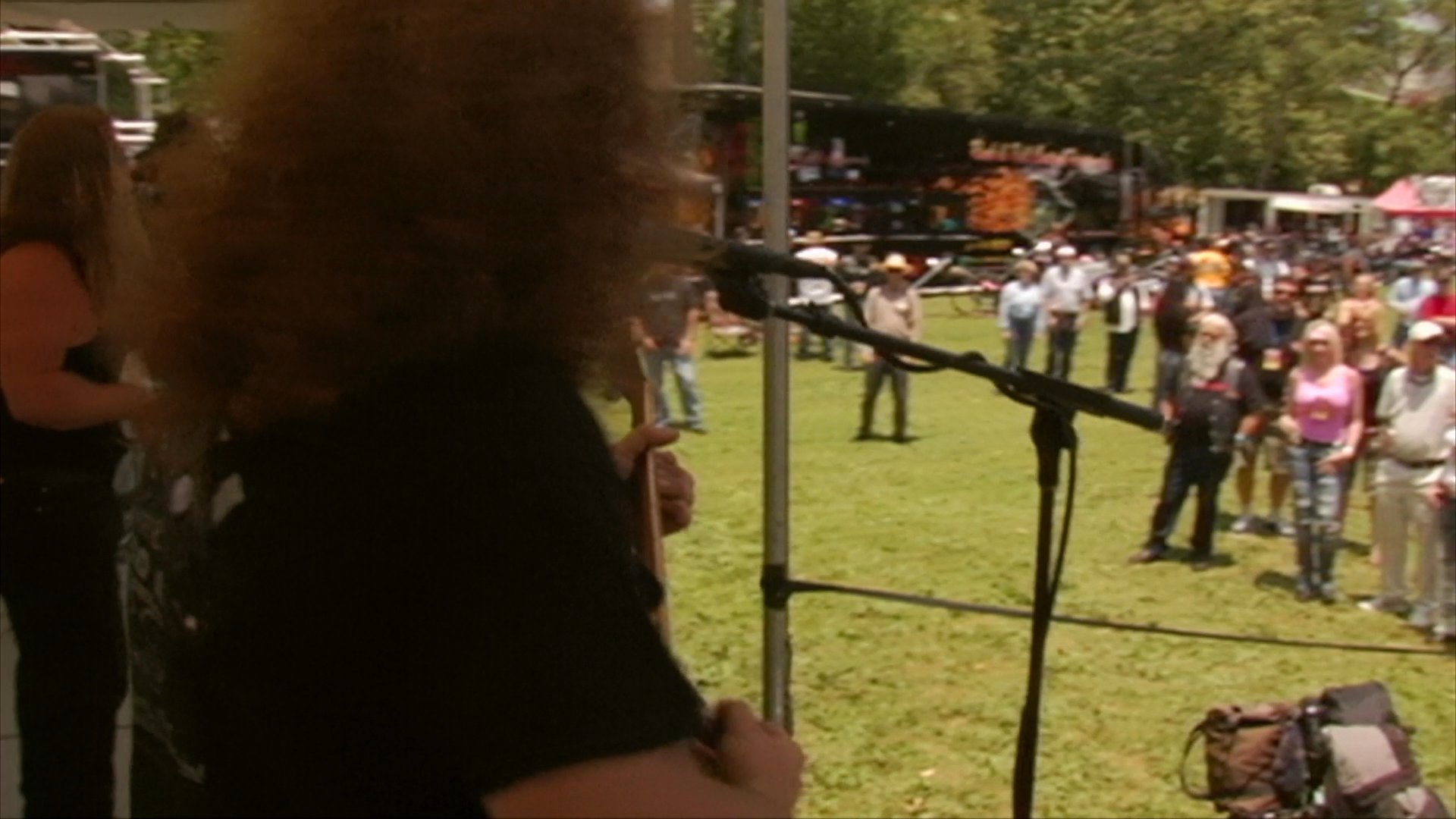 A rock band performs on an outdoor stage for a crowd gathered at a festival event. The musicians, featuring prominent beards and sunglasses, play their instruments while addressing the outdoor audience.