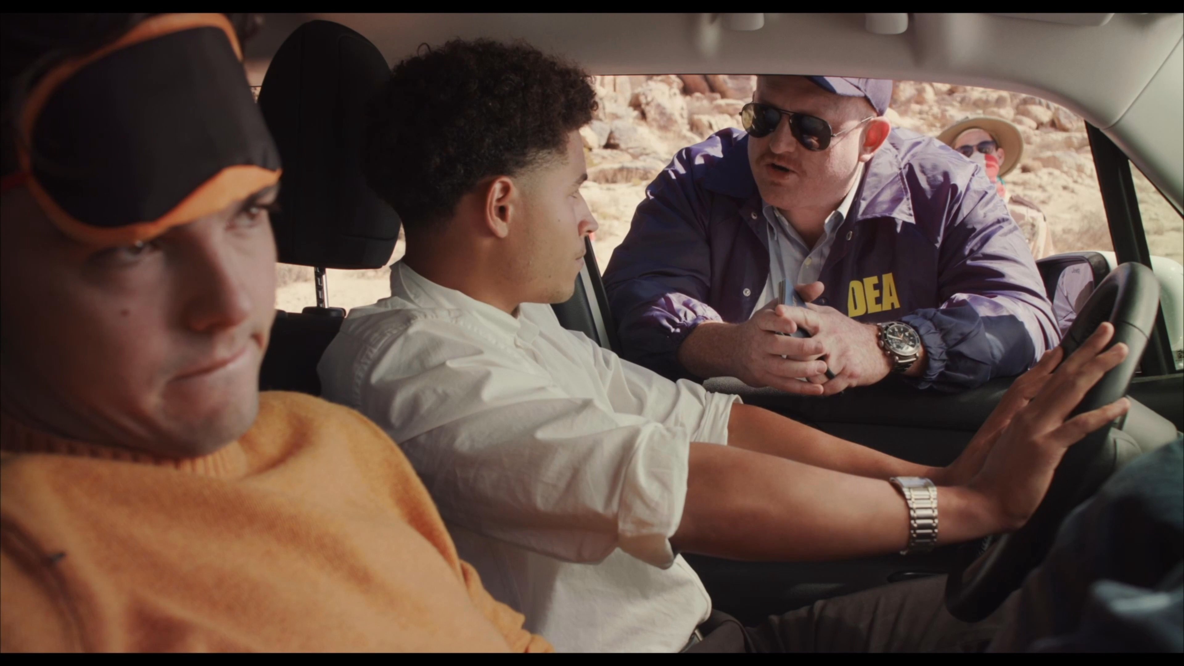 A DEA agent in sunglasses initiates a tense interrogation with two suspects seated inside a vehicle parked in a desert environment.