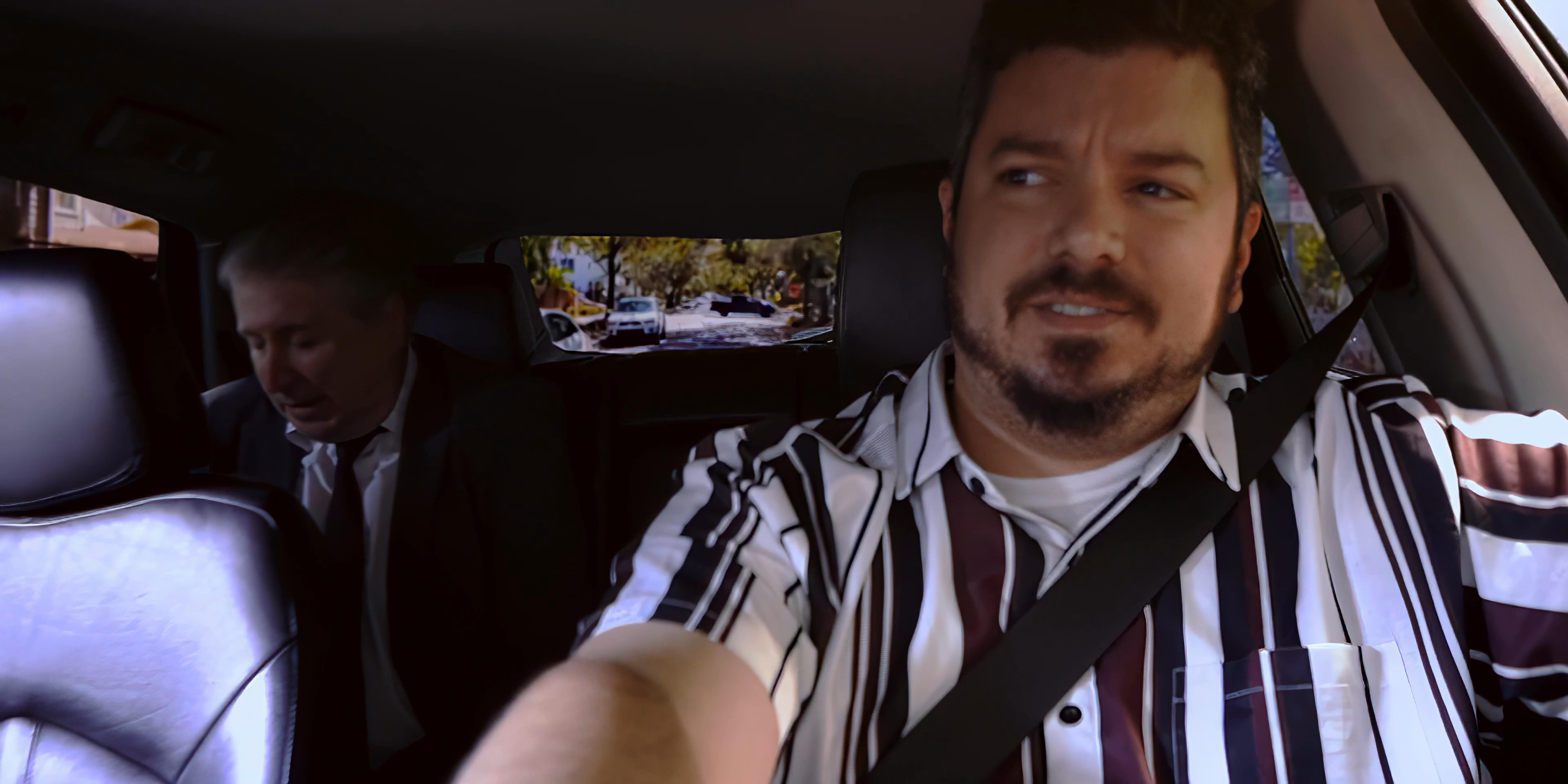 Two men travel through a residential neighborhood, with the passenger observing the surrounding houses while engaged in a conversation with the driver.