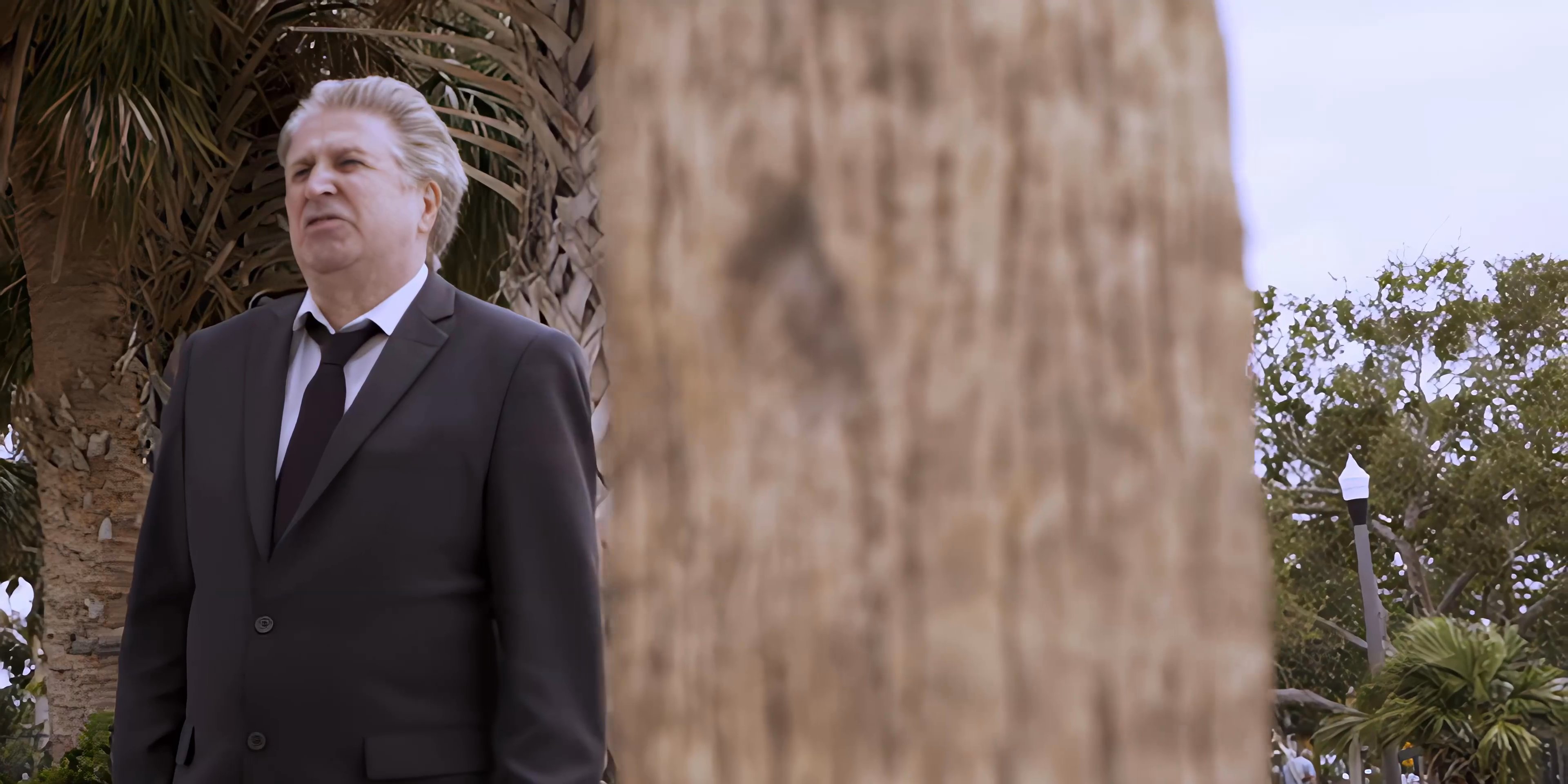 A man wearing a formal suit stands on a beach, looking toward the ocean and palms with a contemplative demeanor. He eventually shifts his gaze and turns slightly, observing his surroundings in a tranquil coastal setting.