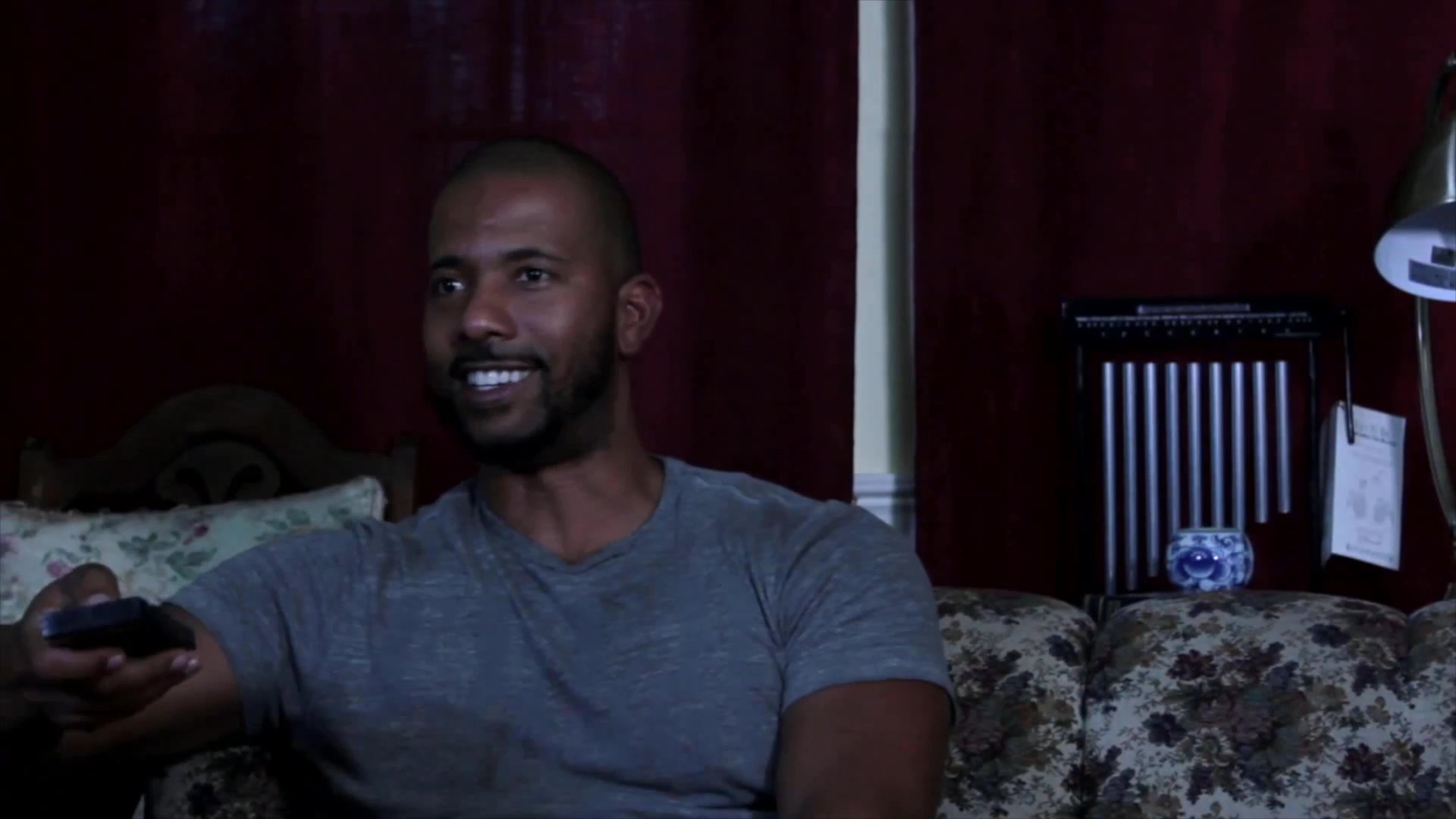 Two men interact in a domestic living room setting, with one individual engaged in a telephone conversation while the other observes.