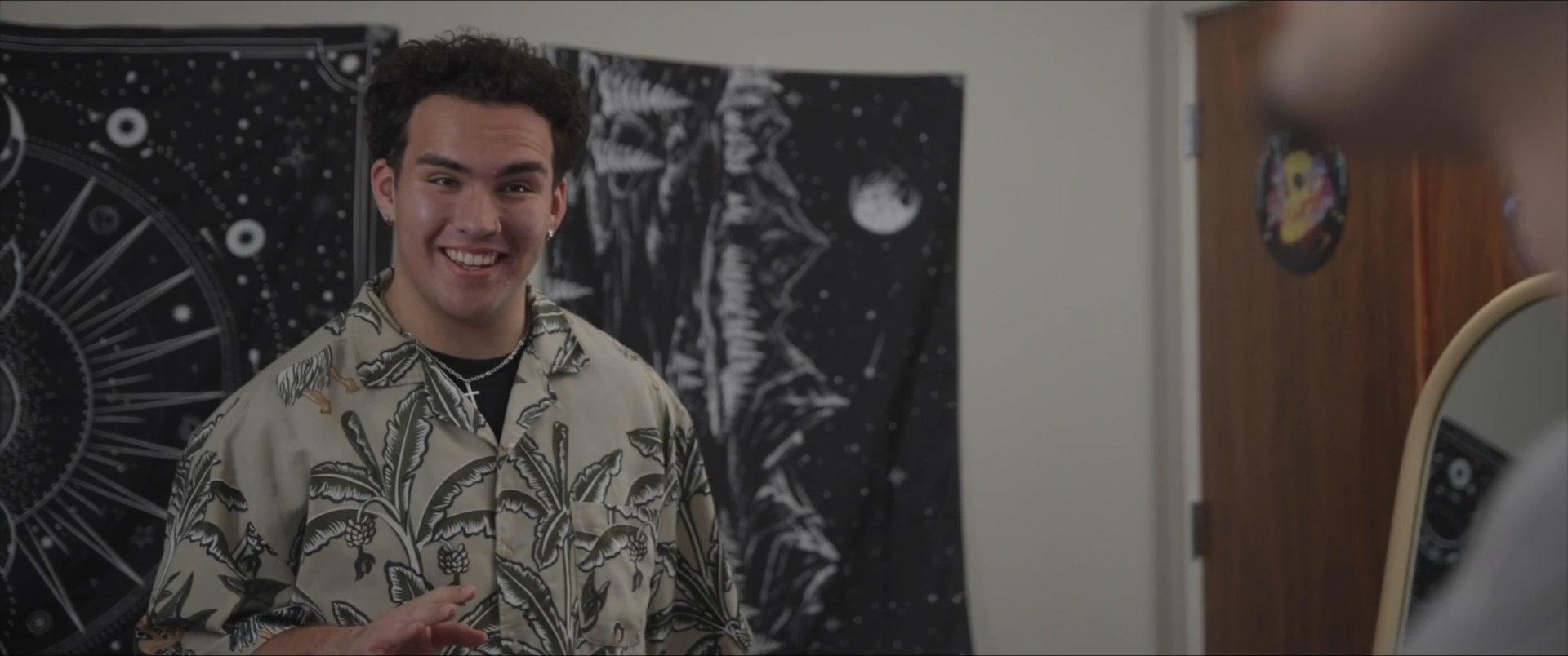 Two young men engage in a lighthearted conversation within a residential interior setting while sharing drinks. They exchange smiles and gestures, reflecting a relaxed and friendly social interaction.