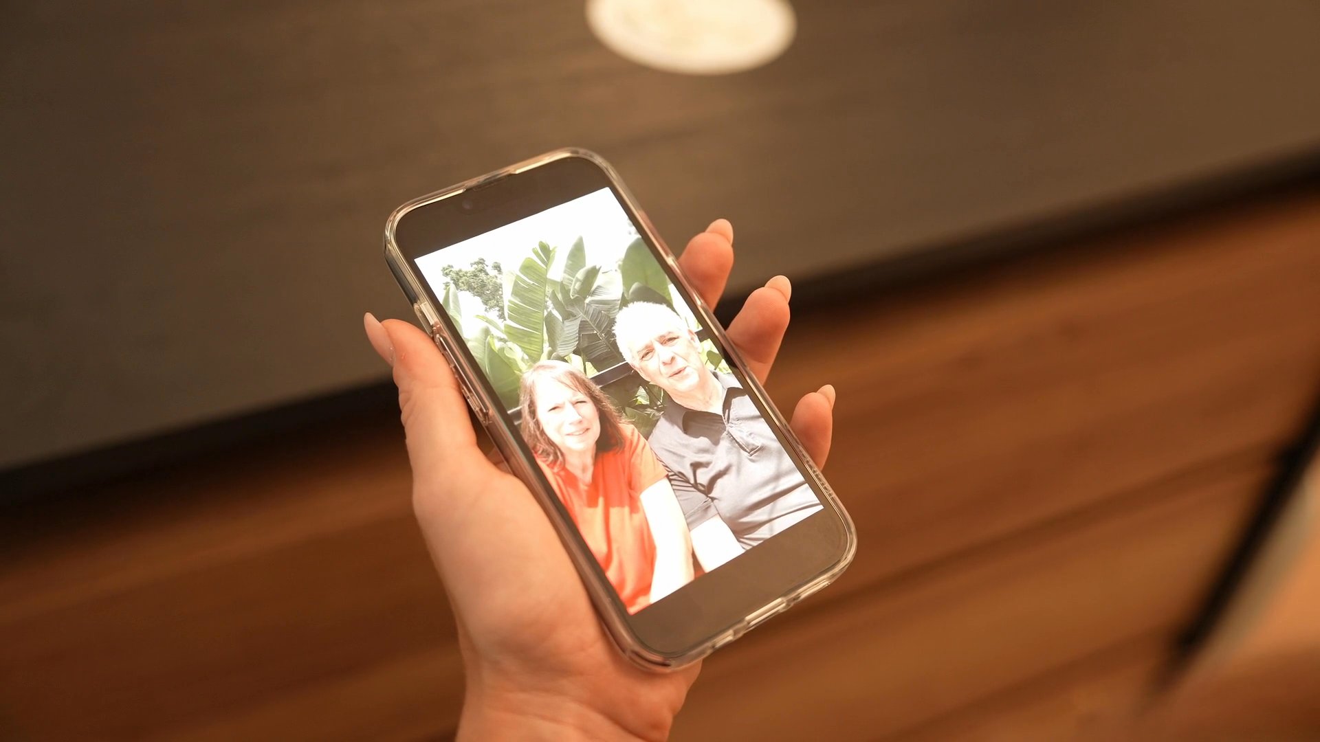 A woman uses a smartphone to view a personal photograph in a residential living space while a man moves through the background. The interaction highlights a moment of digital leisure within a home environment.
