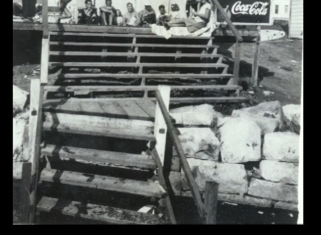 An elderly man recounts his past experiences while viewing archival black-and-white footage of social gatherings at a beachside establishment. The sequence juxtaposes candid moments of youth against a contemporary reflective interview setting.