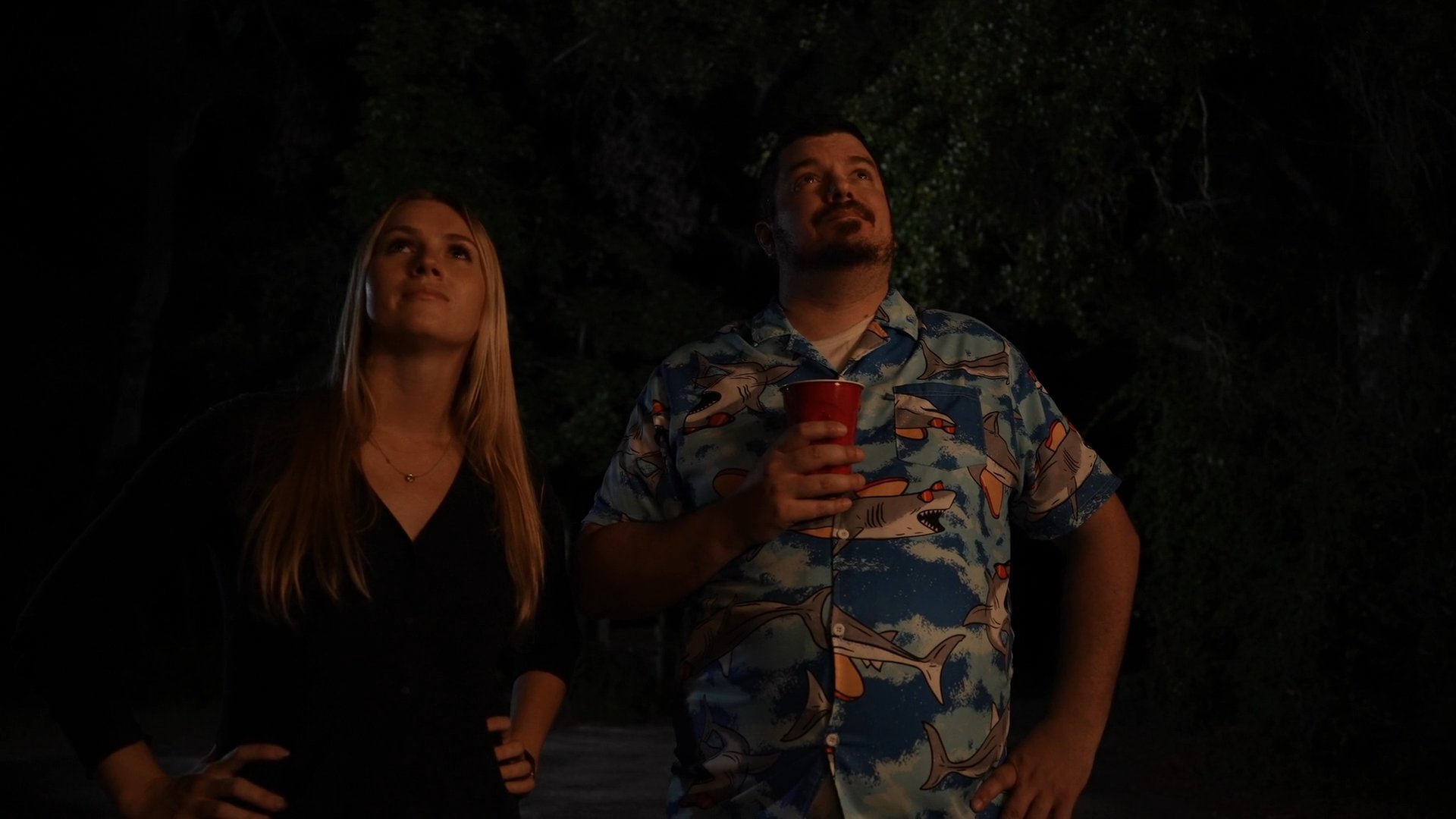 A man and woman stand together in a dark outdoor setting, gazing upward at the night sky filled with stars.