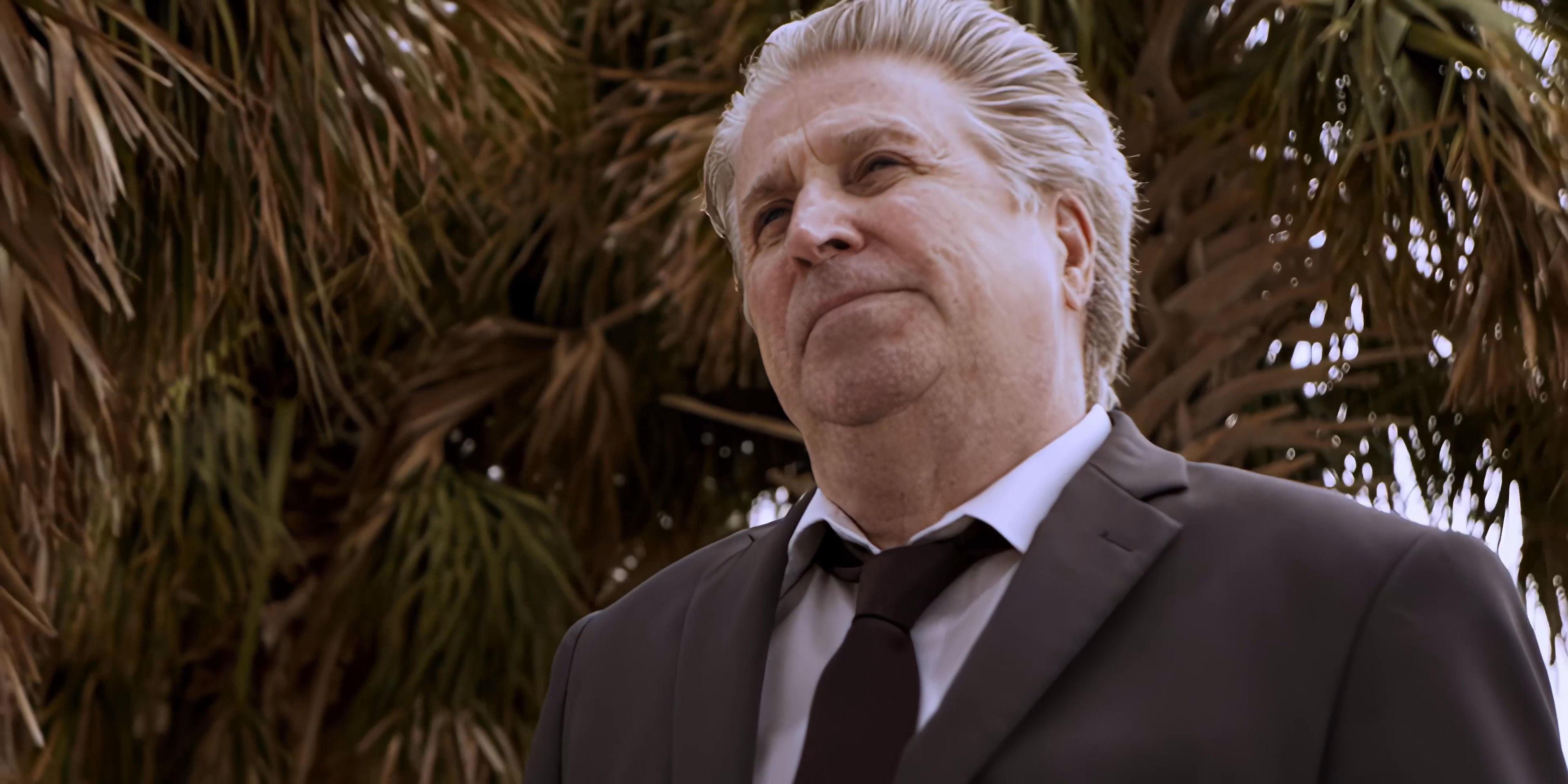 A man in a dark suit stands outdoors beneath palm trees, gazing thoughtfully into the distance.