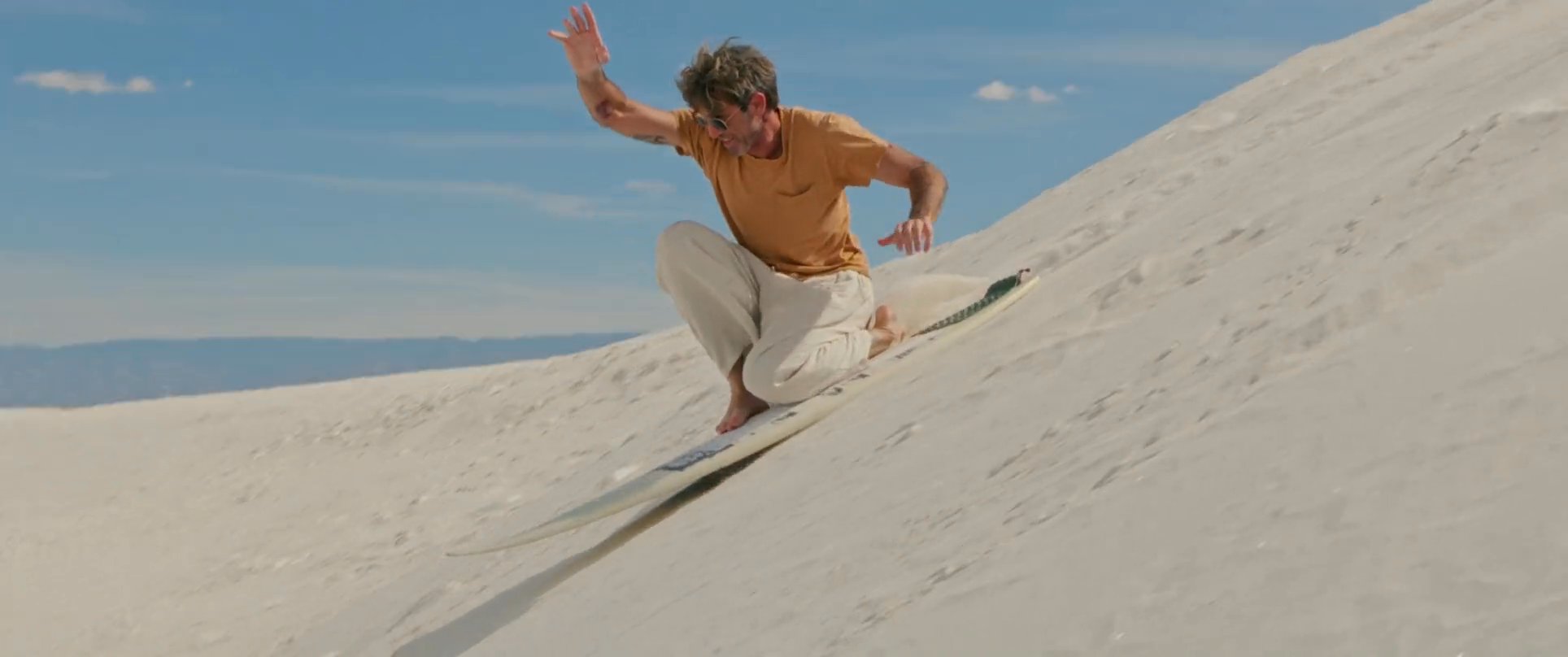 A man navigates a steep sand dune on a board, showcasing various techniques and balancing maneuvers in a desert landscape.