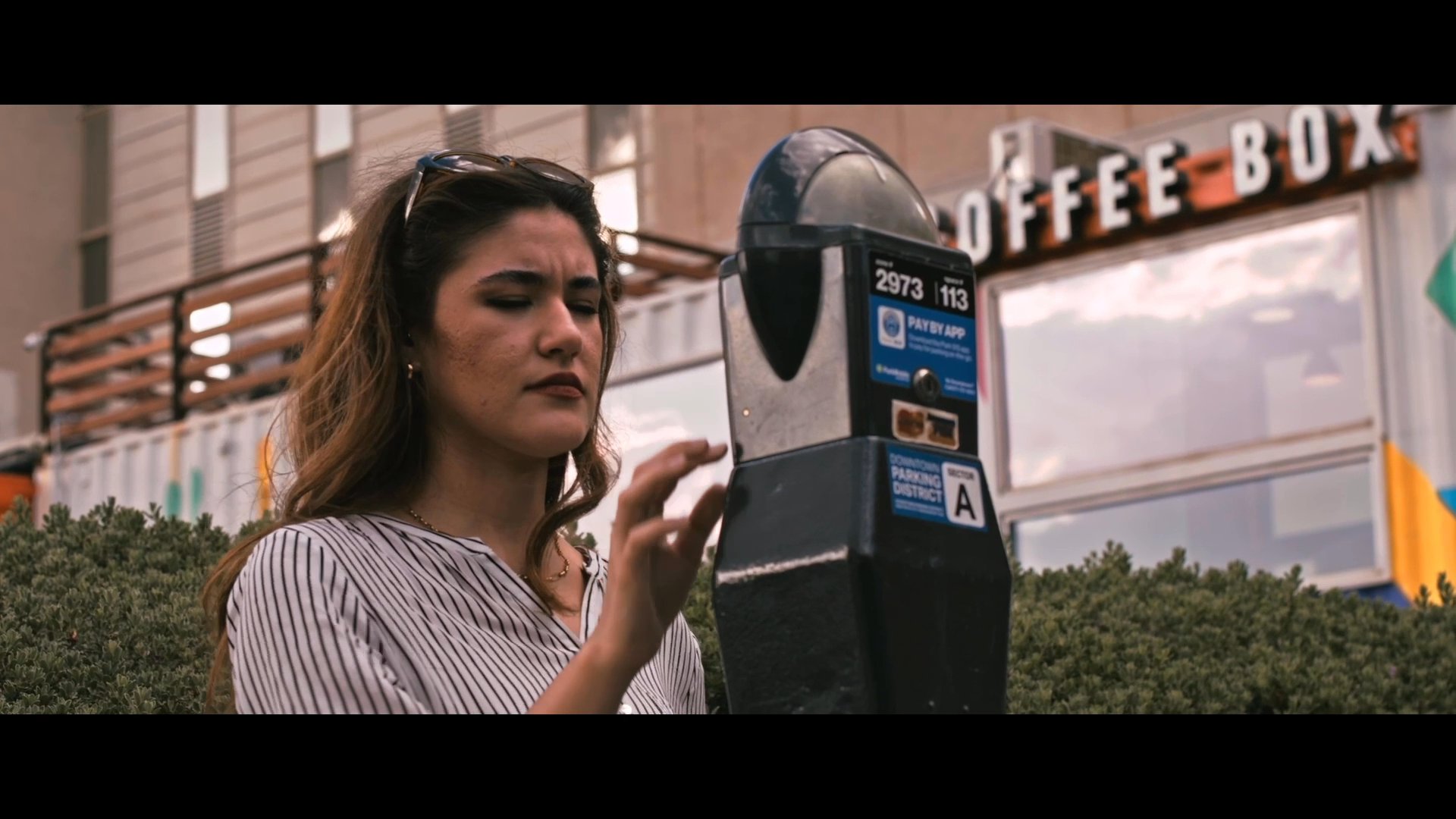 A woman parks her vehicle, pays at a meter, and walks along a city street toward a commercial building. She pauses to observe her surroundings before continuing on foot.