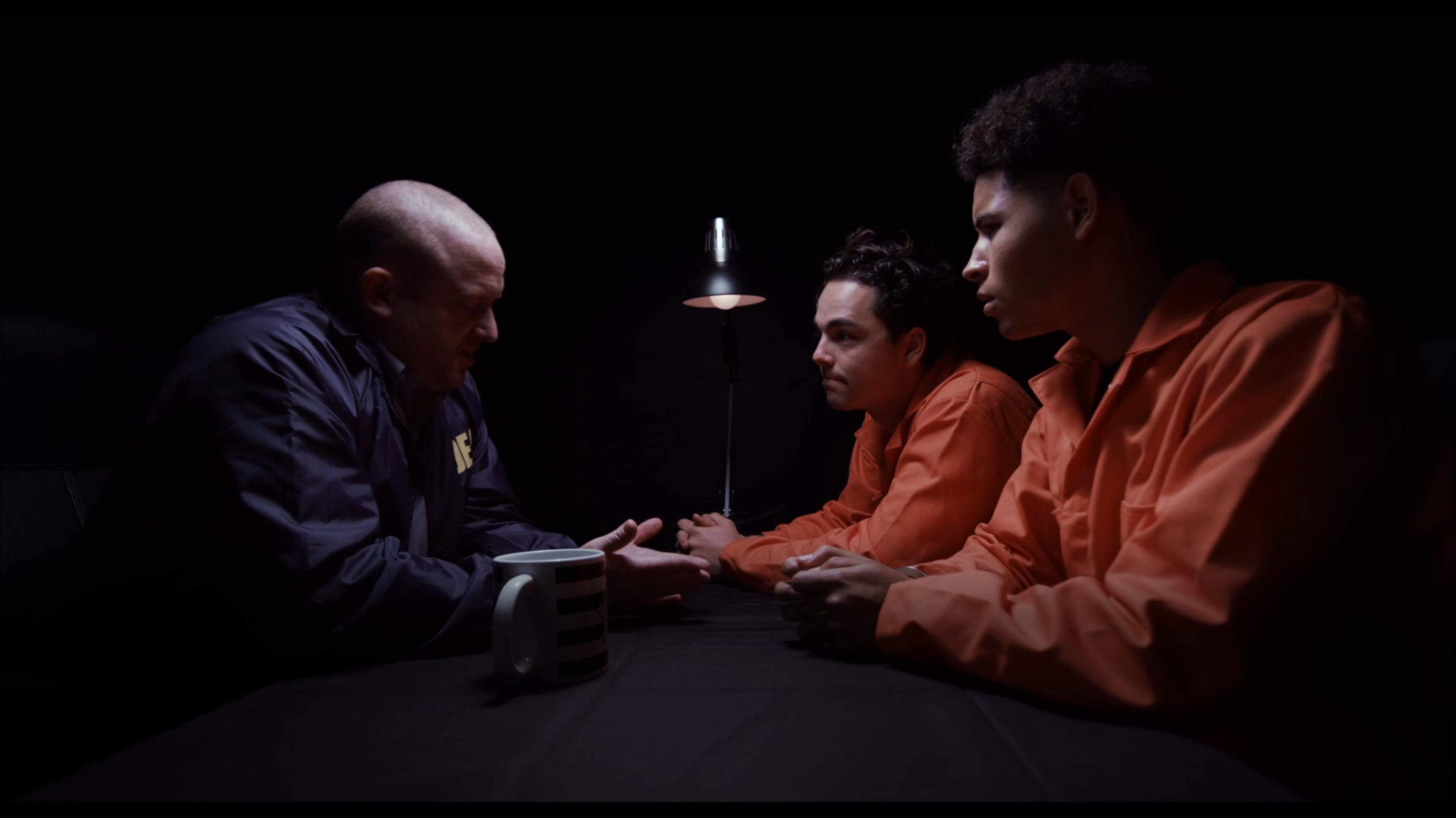 A DEA agent conducts a formal interrogation of a handcuffed suspect in a dimly lit environment. The tense atmosphere defines the exchange as the agent questions the individual regarding their case.