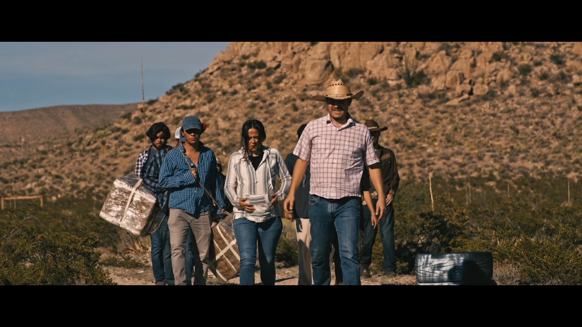 A group of individuals travels across an arid, sunlit desert landscape. The journey highlights the physical exertion and collective movement of the group through challenging terrain.