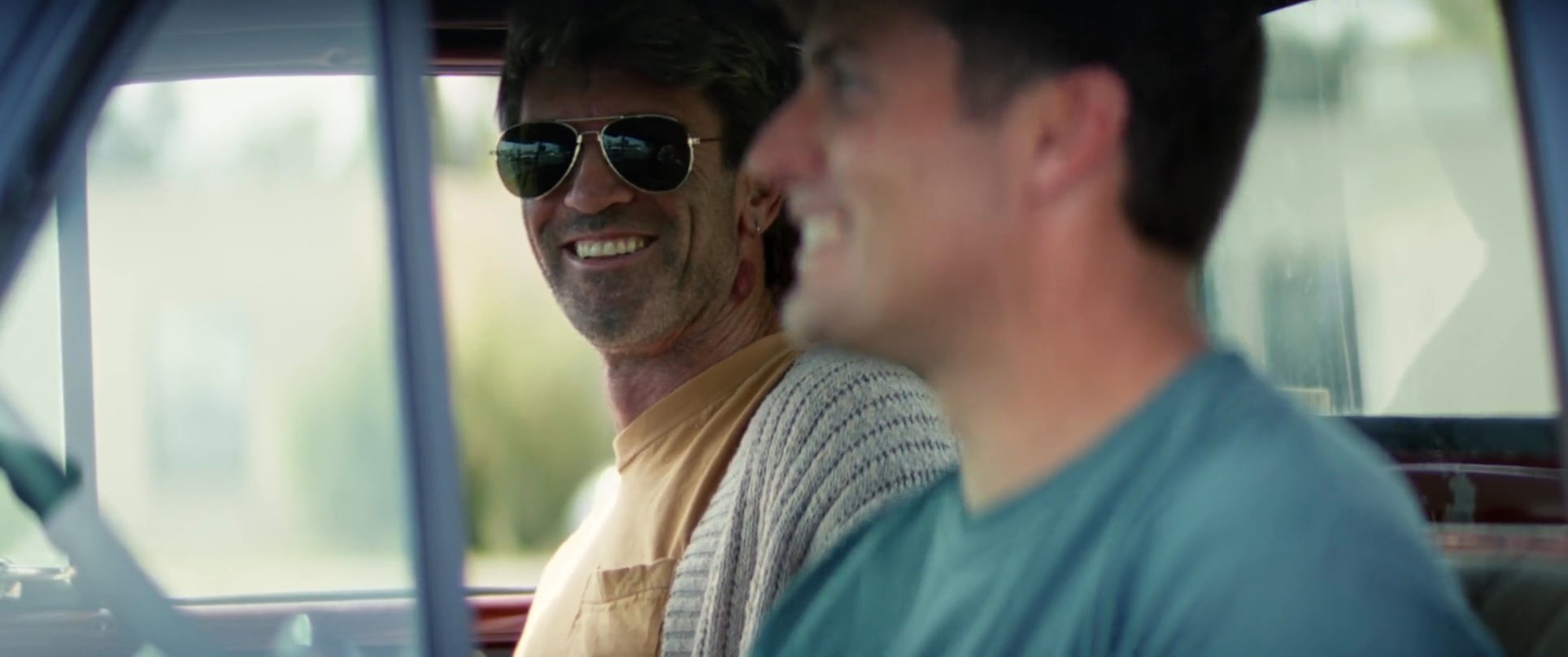 Two men laugh and engage in a lighthearted conversation while seated inside a vintage truck.