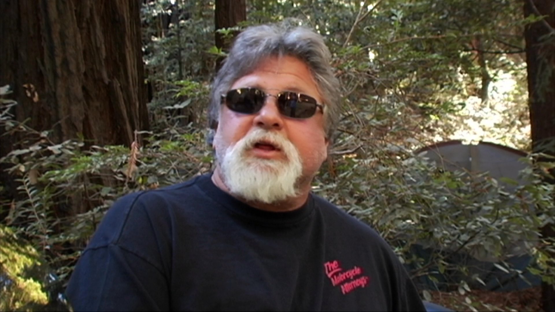 Participants gather at an outdoor motorcycle rally featuring a large collection of bikes to support a charity cause. An organizer shares details about the event while attendees and supporters socialize in a park setting.