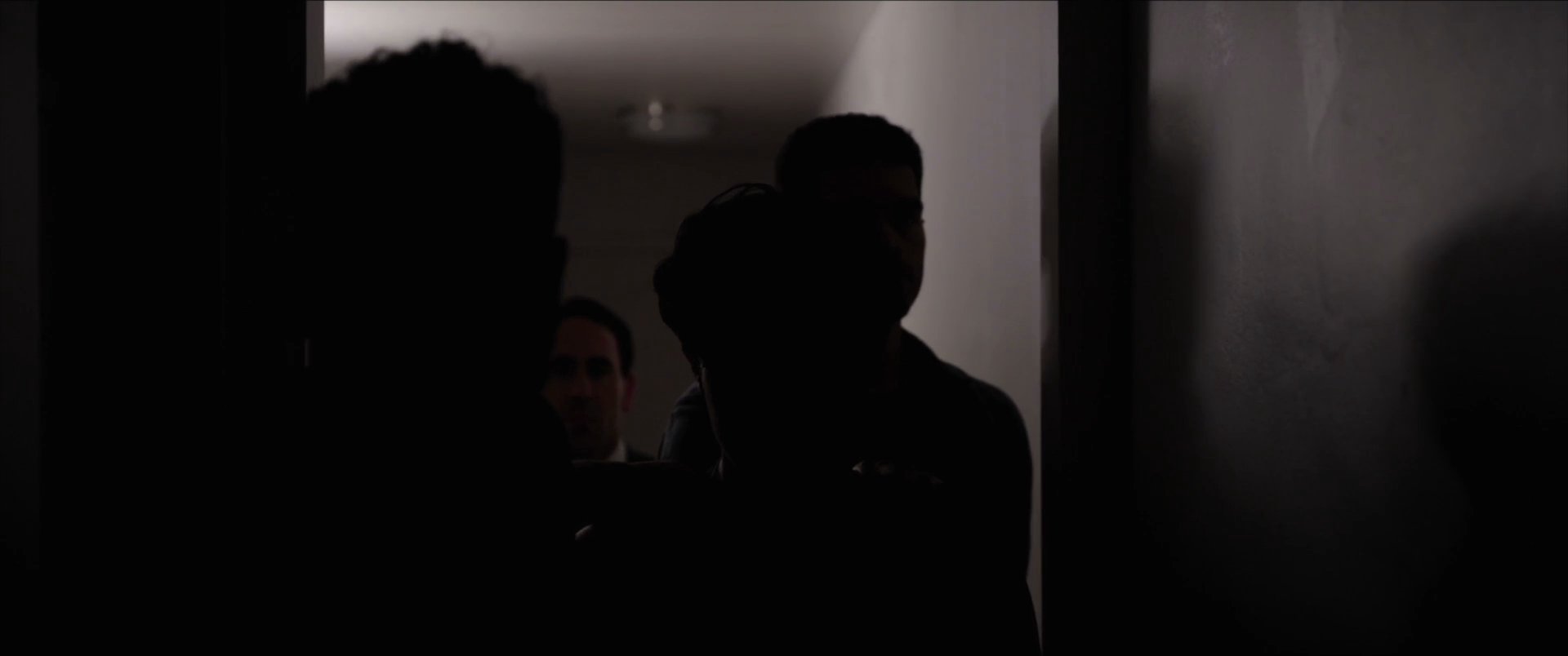 A group of men navigates a dimly lit residential hallway, transitioning from light into deepening shadow. The sequence emphasizes a tense, cinematic atmosphere as the figures move collectively through the narrow passage.