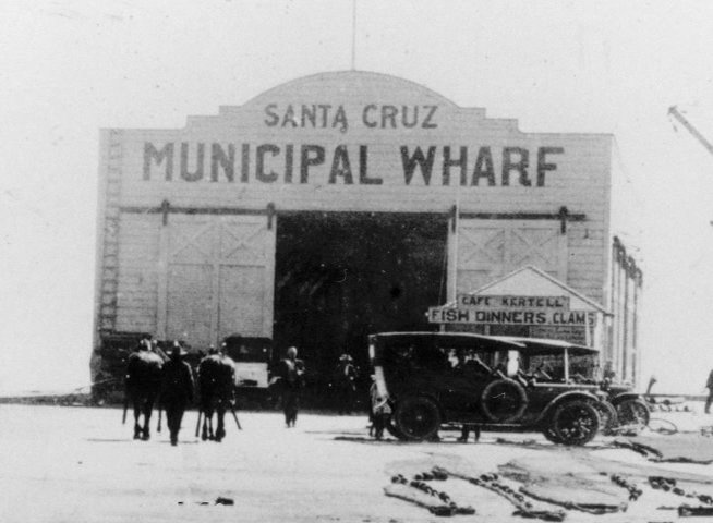 Pedestrians and vintage automobiles navigate the Santa Cruz Municipal Wharf in early 20th-century California. The sequence concludes with a group of early surfers posing with their boards on the beach.