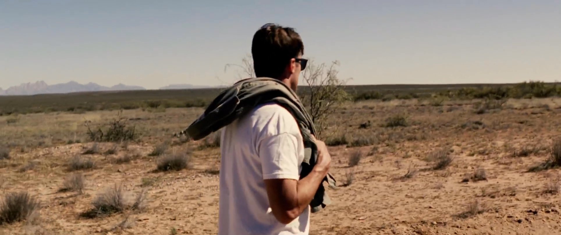 A man in a white t-shirt and sunglasses walks alone along a desolate dirt road in an arid desert environment, carrying a jacket over his shoulder.