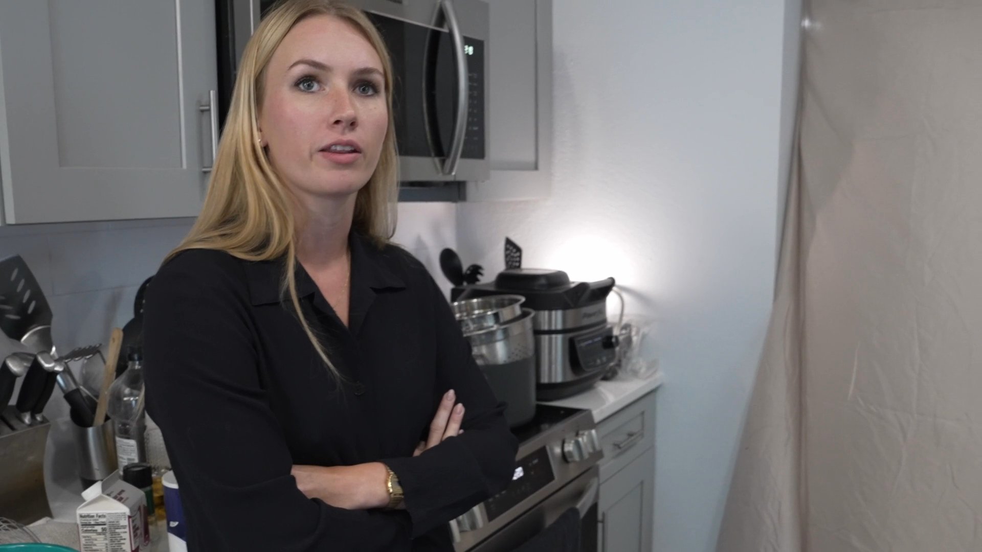 A woman meticulously records information in a notebook while a man in a patterned shirt engages her in a tense kitchen conversation.