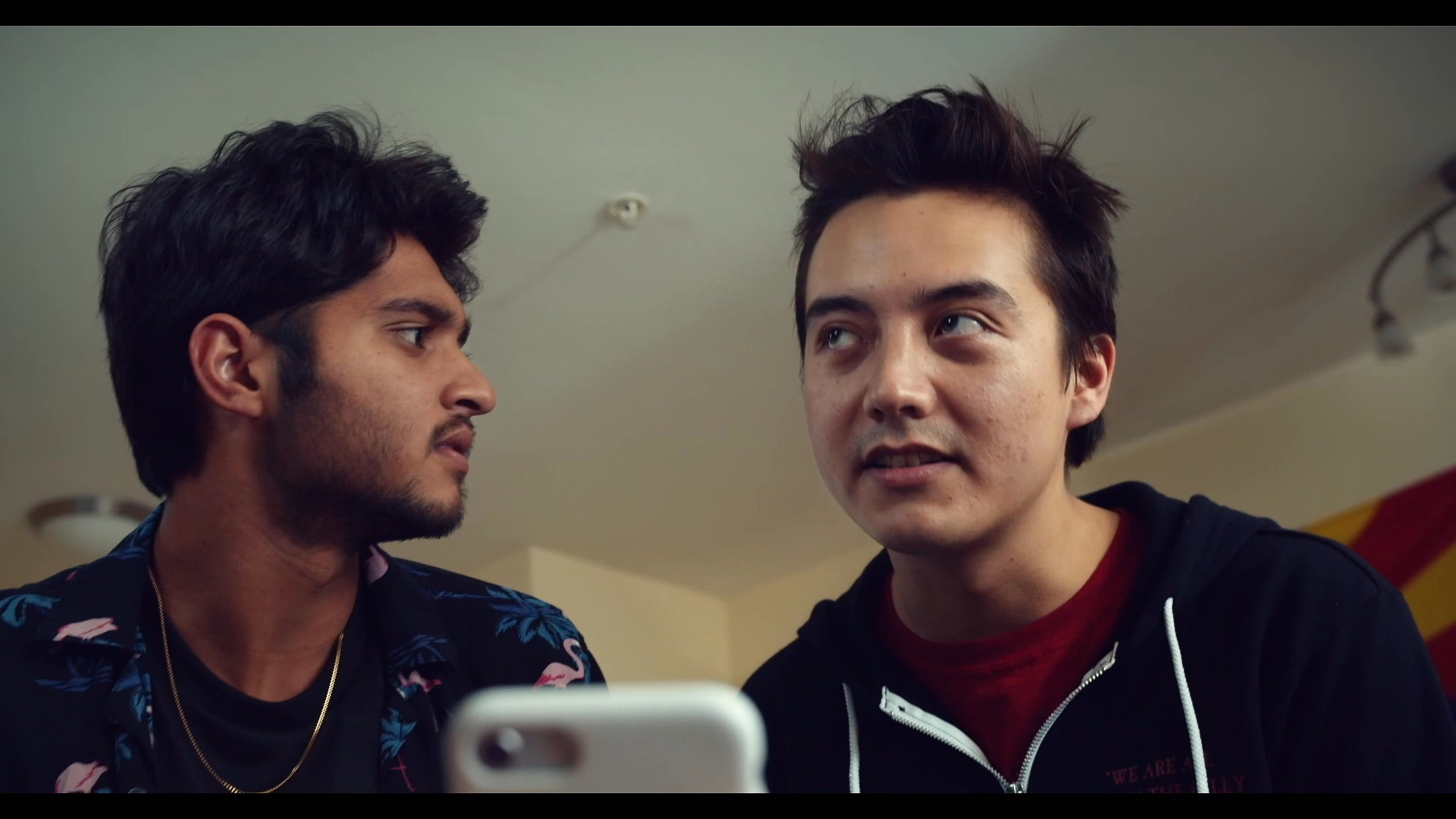 Two friends review a social media post showing a newborn baby on a smartphone before moving into a kitchen setting to discuss the news with another person.