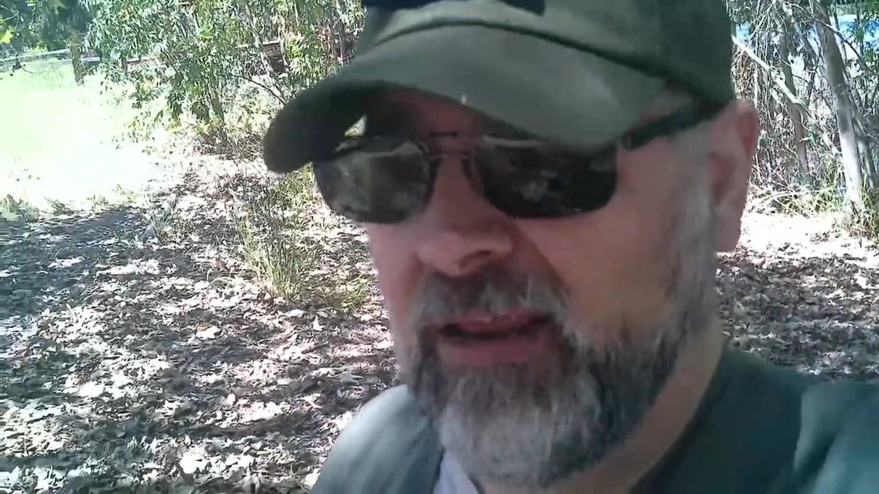 A man with a beard and tactical cap speaks directly to the camera while situated in a sunlit, wooded outdoor environment.
