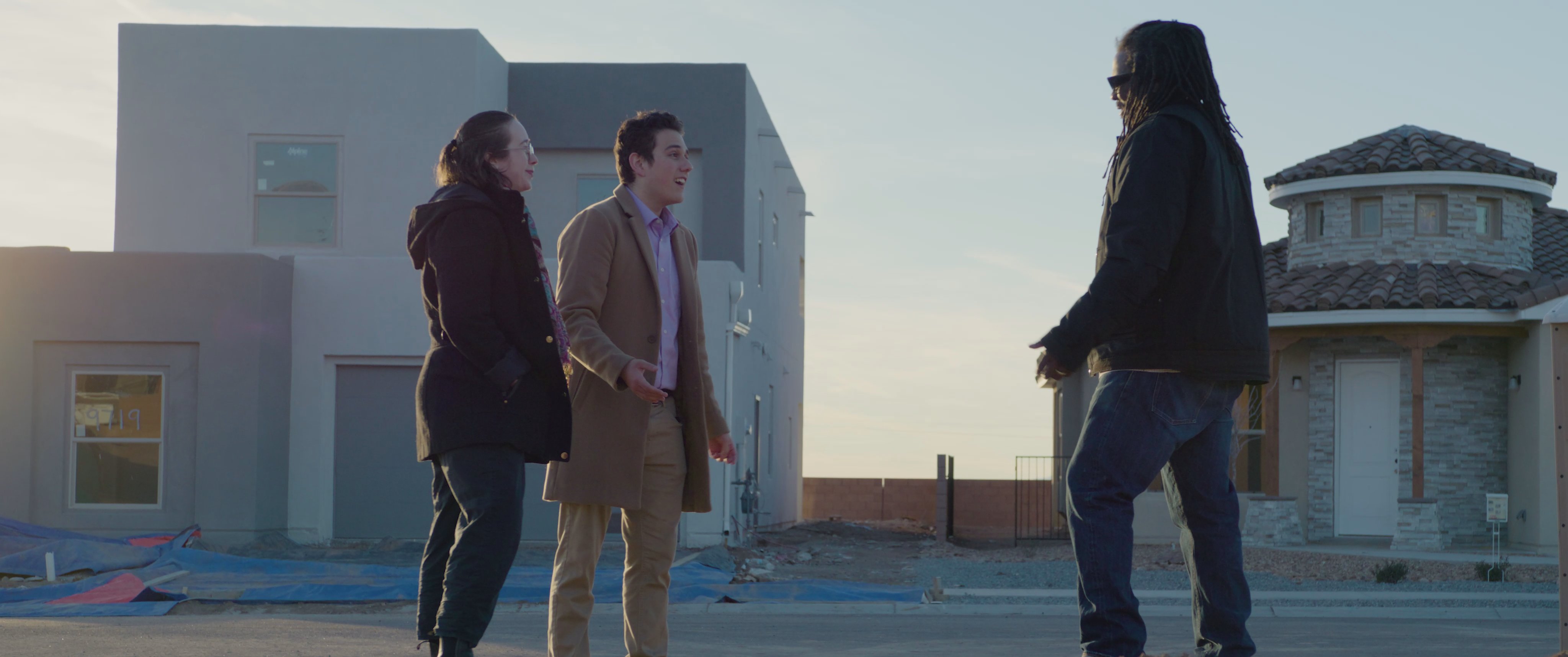 Prospective clients meet with a contact at a new residential construction site to discuss property details. The group engages in conversation before a woman emerges from the home to join the interaction.