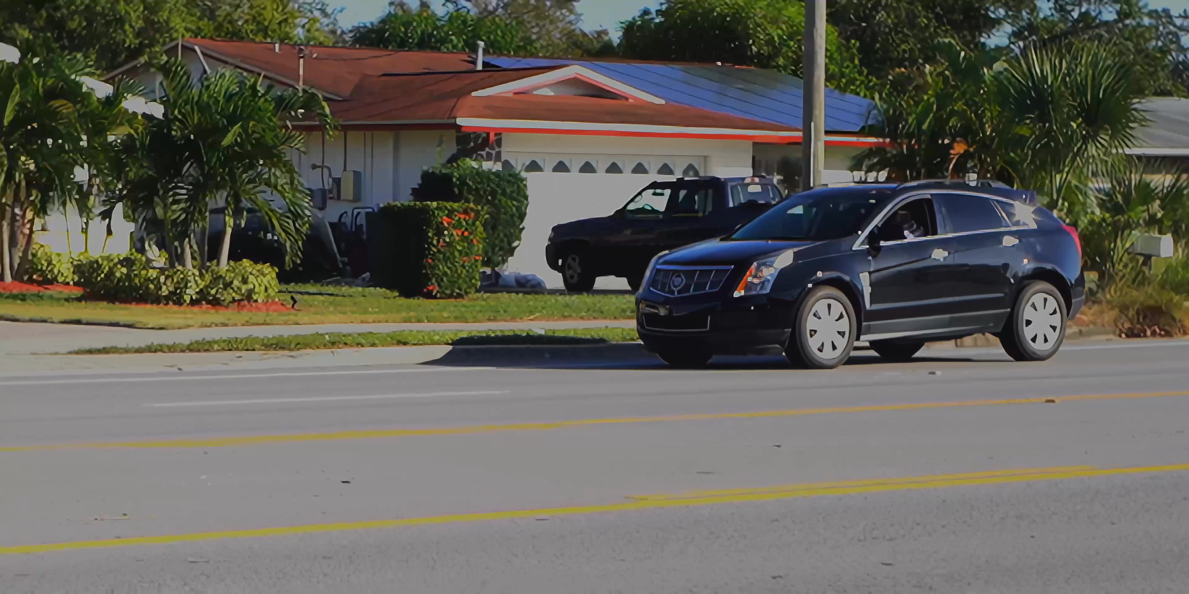 A vehicle traverses a suburban neighborhood, passing residential homes and parked cars under a clear, sunlit sky. The perspective transitions from a roadway view to a close-up of a dark SUV parked in a driveway.