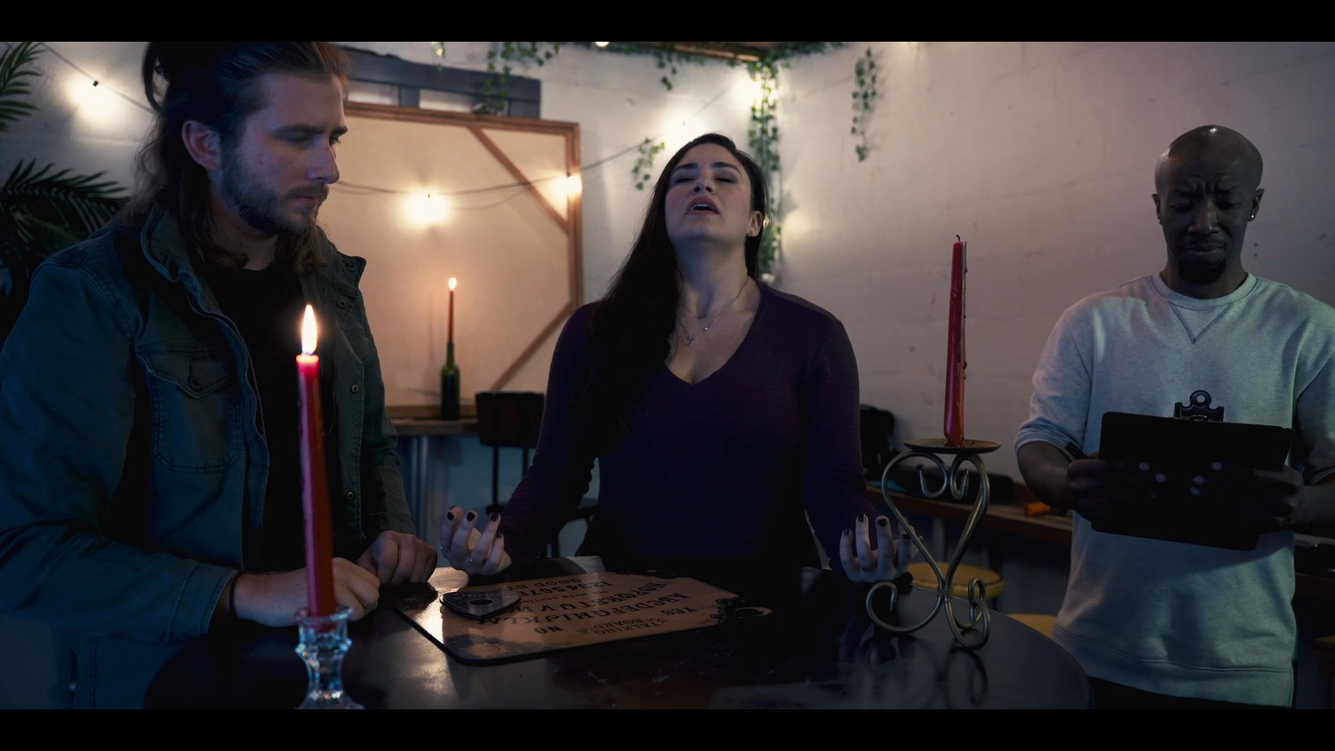 Three individuals participate in a candlelight seance, interacting with a ouija board while one person documents the session. The atmosphere is tense as they attempt to make contact with the supernatural.