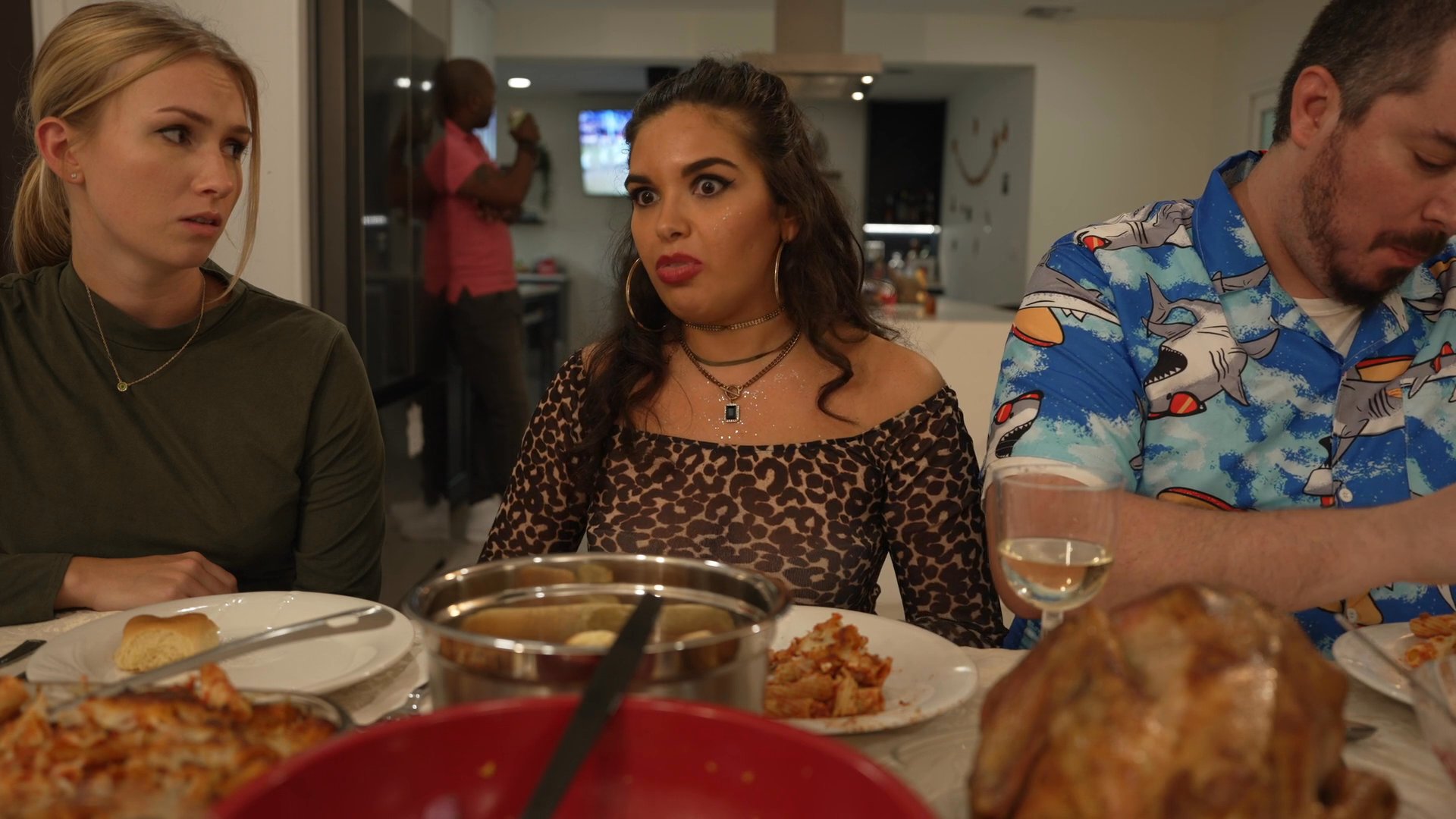 A group of friends engages in an animated discussion while seated at a dining table. The scene highlights interpersonal dynamics and social interaction during a meal.