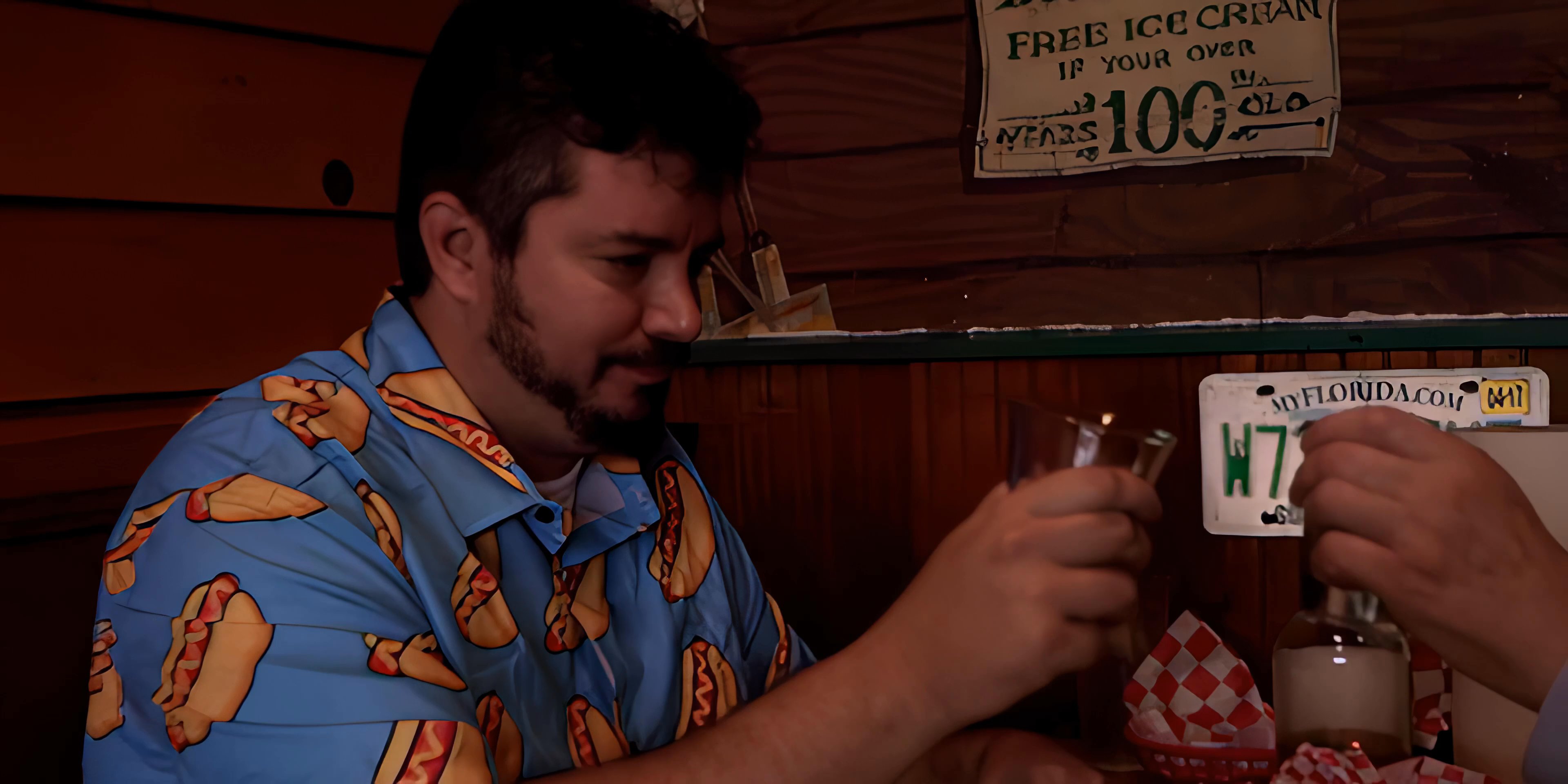 Two individuals, one dressed casually and the other in a dark suit, engage in a conversation while sharing a toast inside a wood-paneled diner booth.