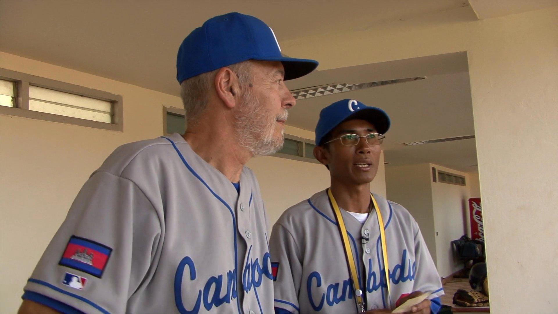 Members of the Cambodia baseball team discuss strategy in the dugout before their match against the Philippines. The scene shifts to an on-field perspective where the players execute drills on the diamond.