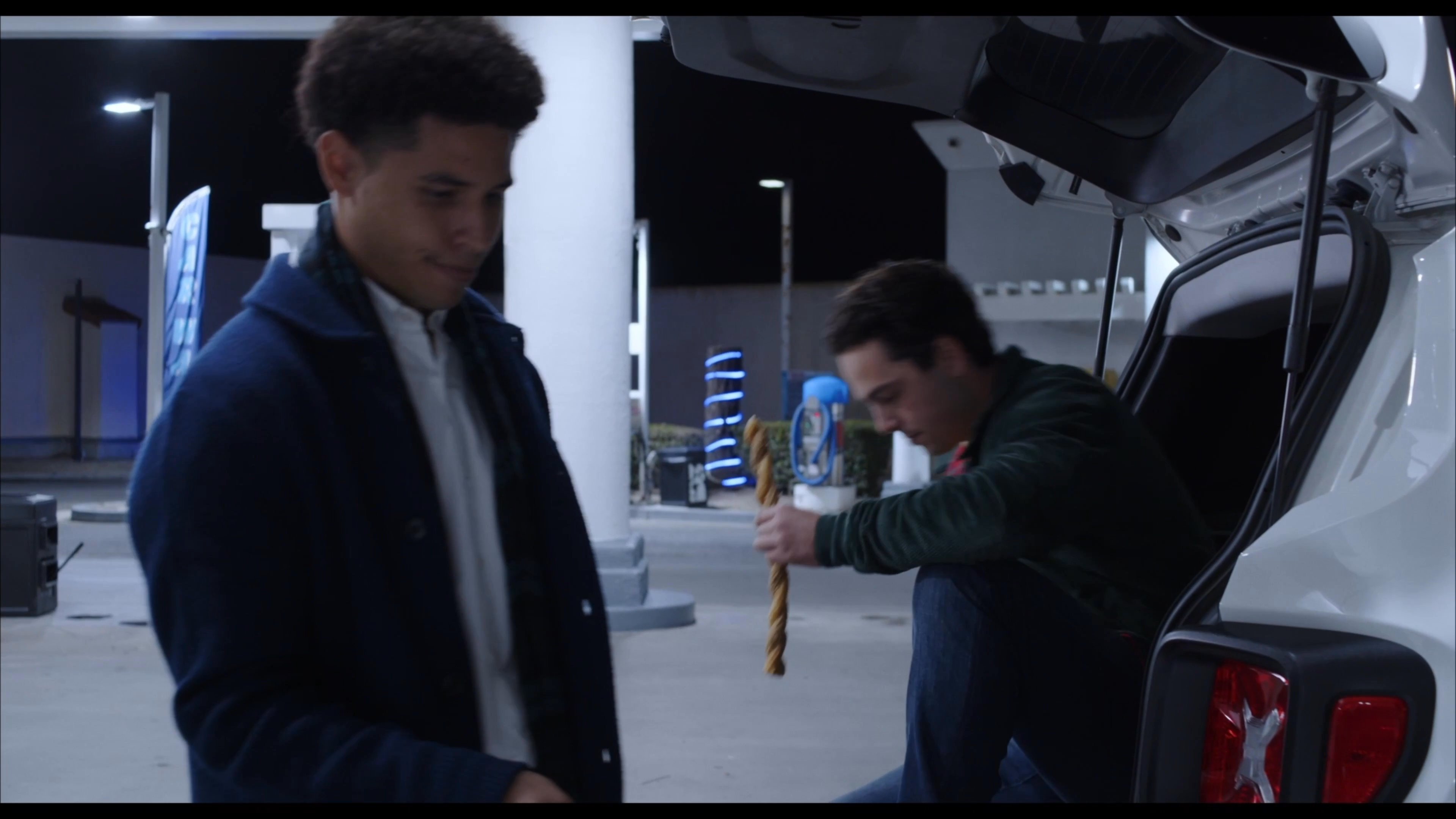 Two young men socialize and consume snacks while sitting at the rear of an open vehicle at a gas station at night.