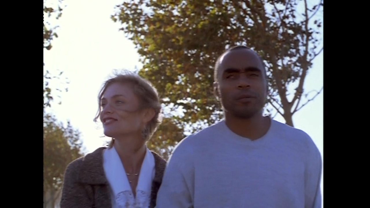 A man and a woman walk arm-in-arm along a paved pathway in a park setting, engaged in conversation.