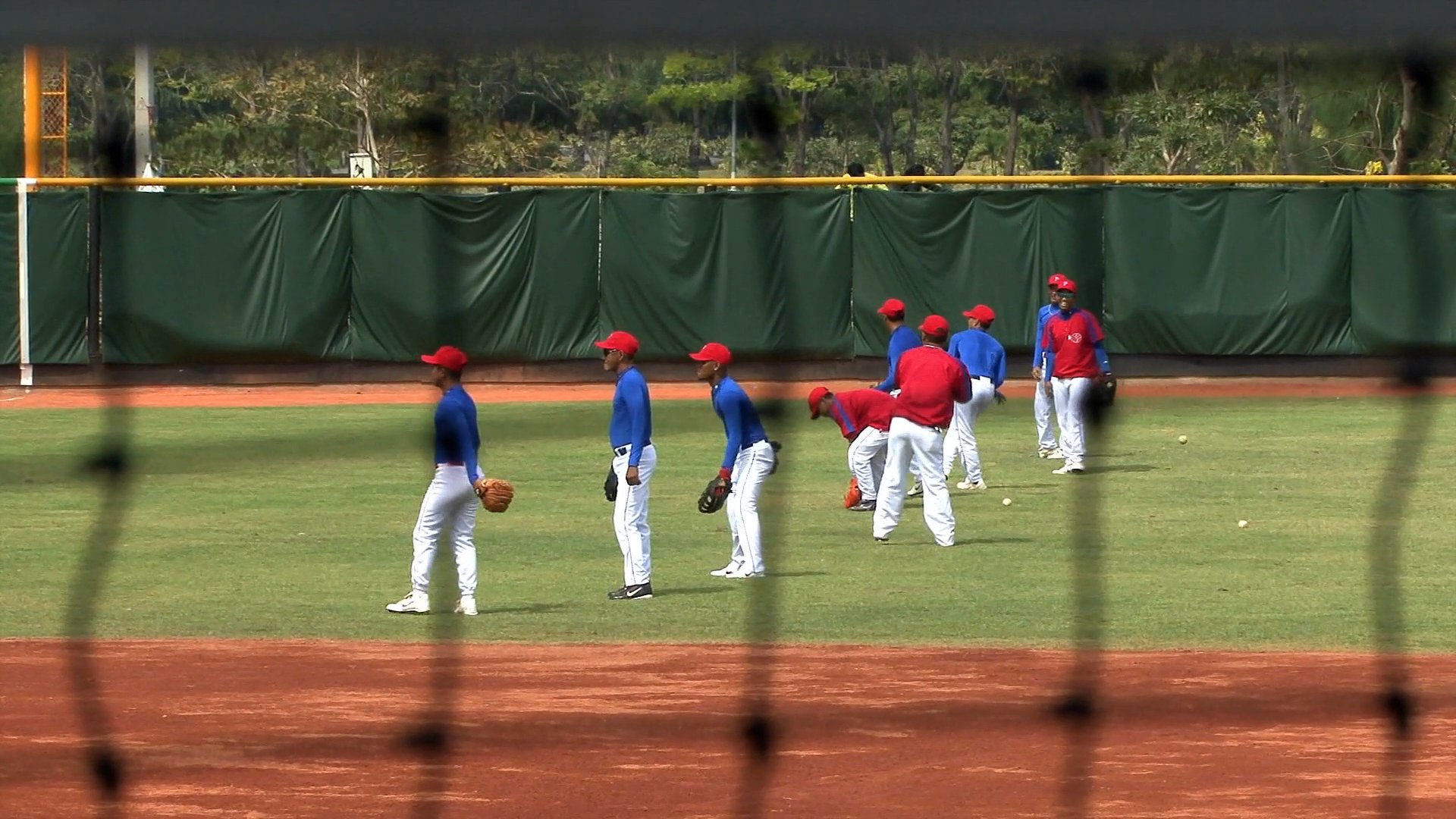 Two team members discuss training strategies in the dugout while teammates practice fielding and batting drills on the baseball field.