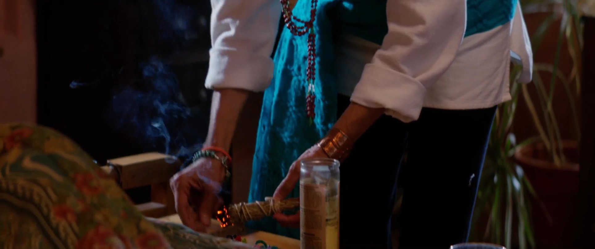 An elder facilitates a traditional smudging ceremony, burning sage to initiate a spiritual transition. Observers sit in quiet contemplation while incense smoke permeates the room.