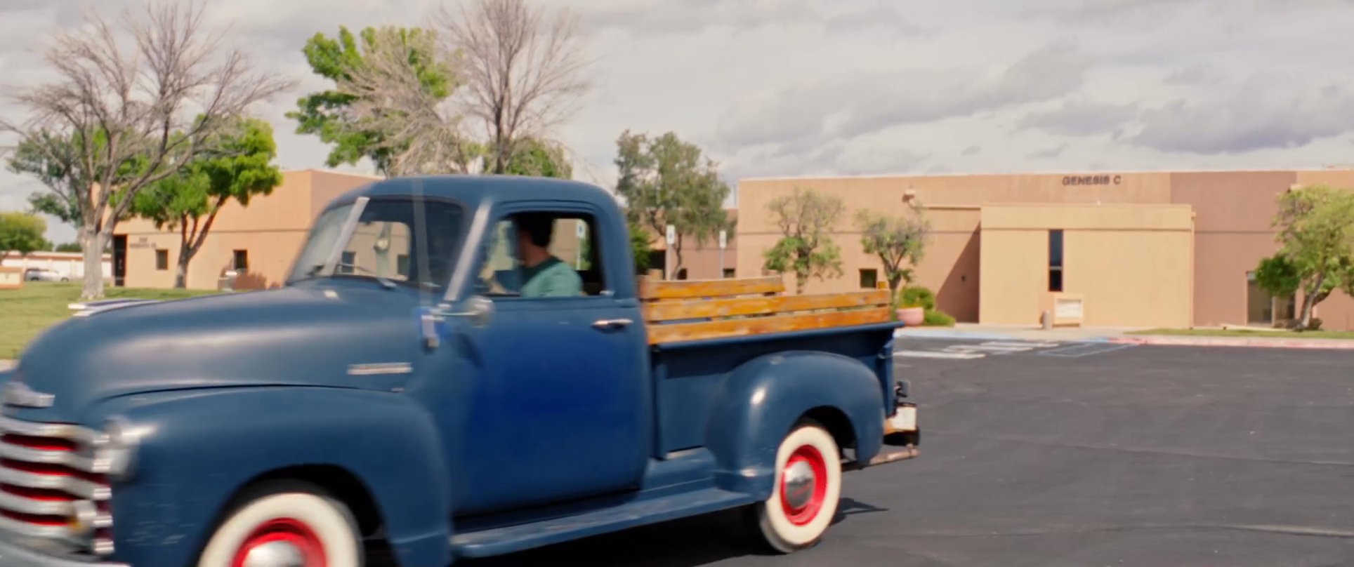 Two men travel through an arid, desert landscape in a classic blue pickup truck, transitioning from an urban parking lot to an open highway.
