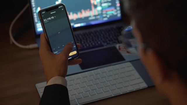 A trader performing financial analysis of cryptocurrency using a phone app and laptop.