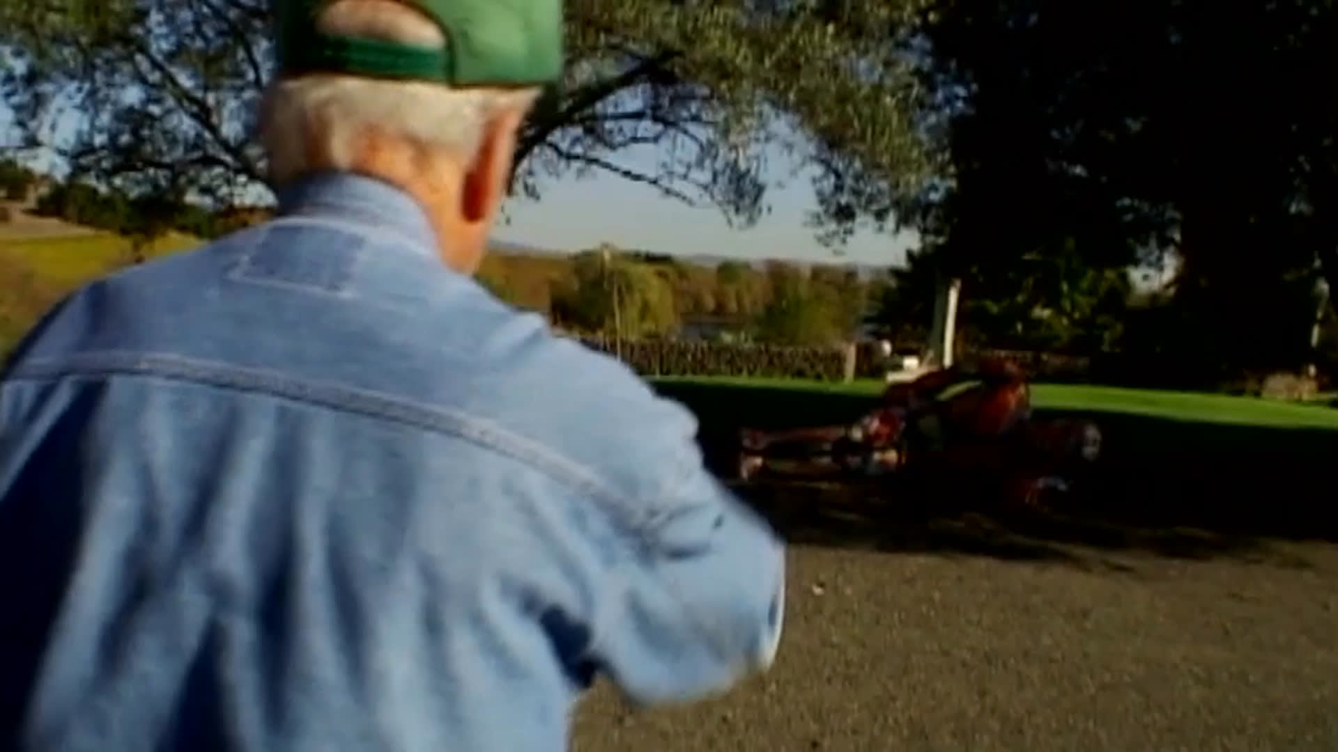 An older man in a green cap and denim shirt walks through a landscaped outdoor setting, passing colorful sculptures and other artworks. He discusses the appeal of a delicate chair and a concrete piece, with a stone wall and river visible nearby.