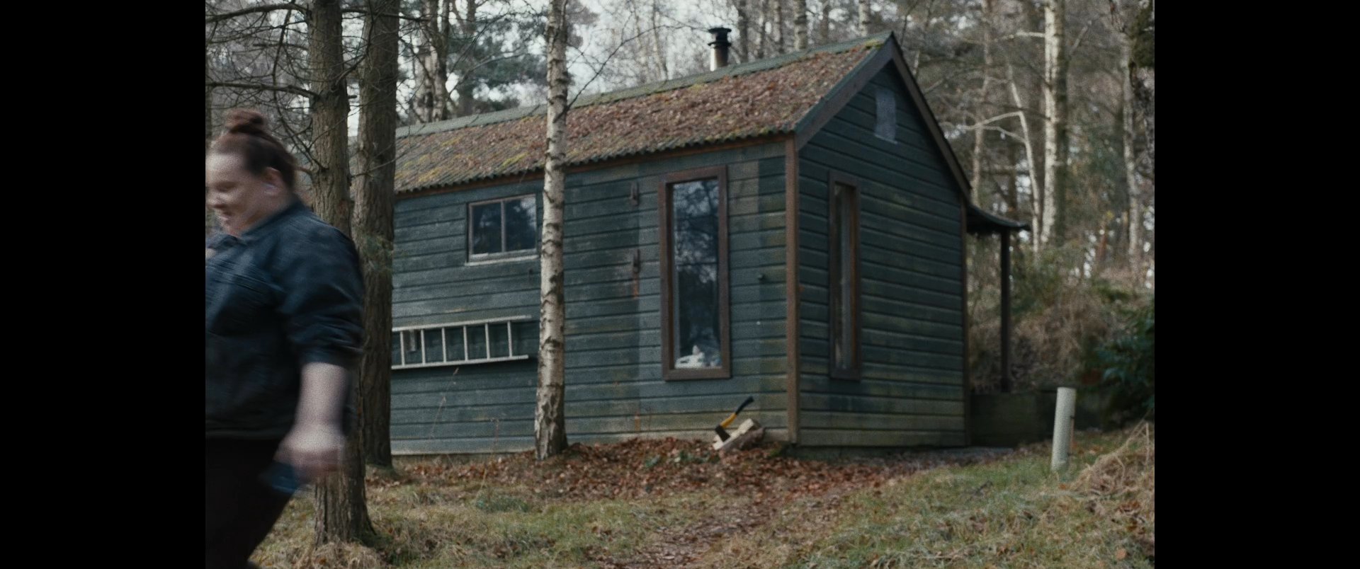 A woman departs a secluded cabin in the woods and walks along a forest path while a person records her with a smartphone.